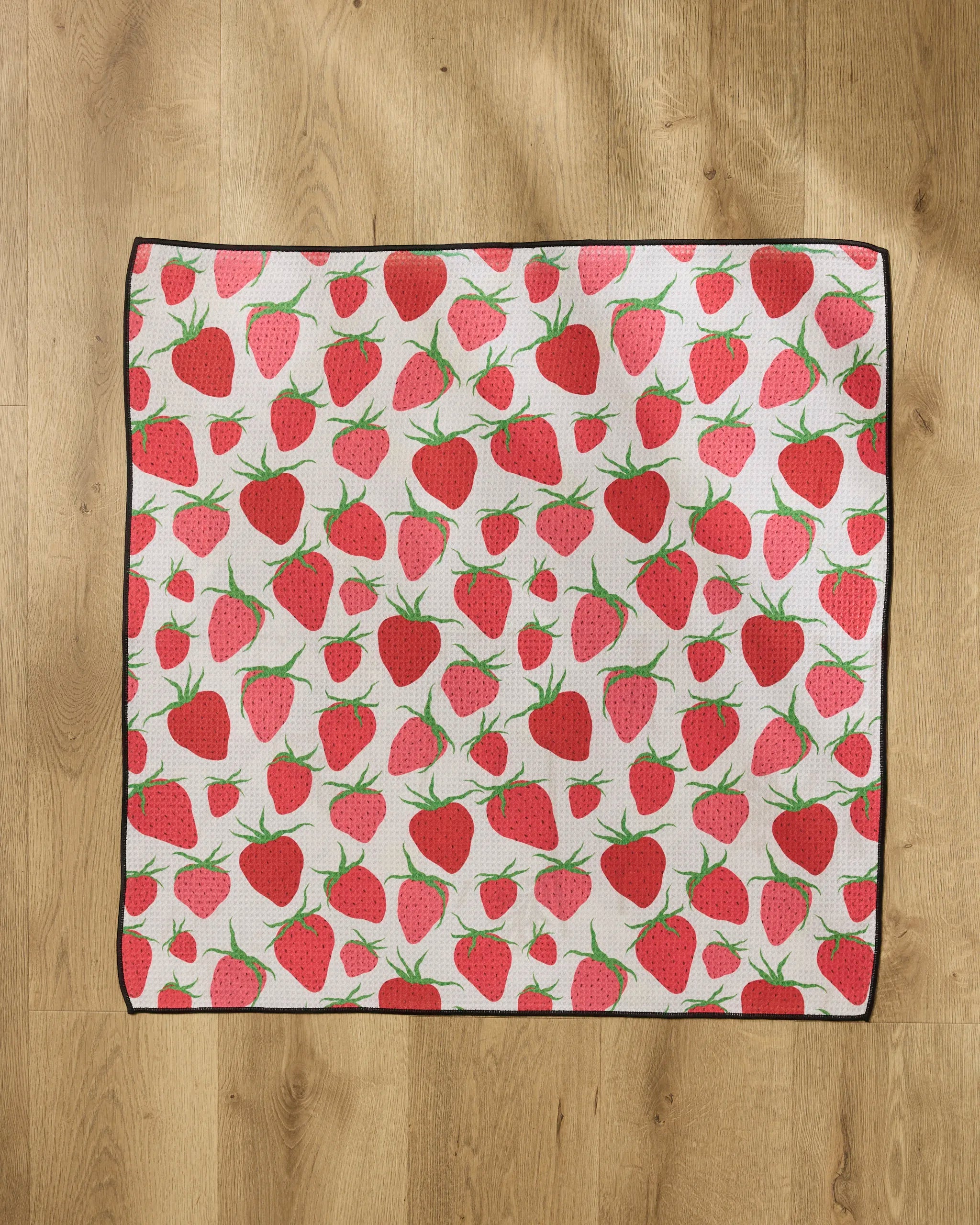 The Geometry Strawberry Sniff dog towel, made from recycled materials, features a white background with red strawberries and green stems, a black border, and is laid flat on a light wooden floor.