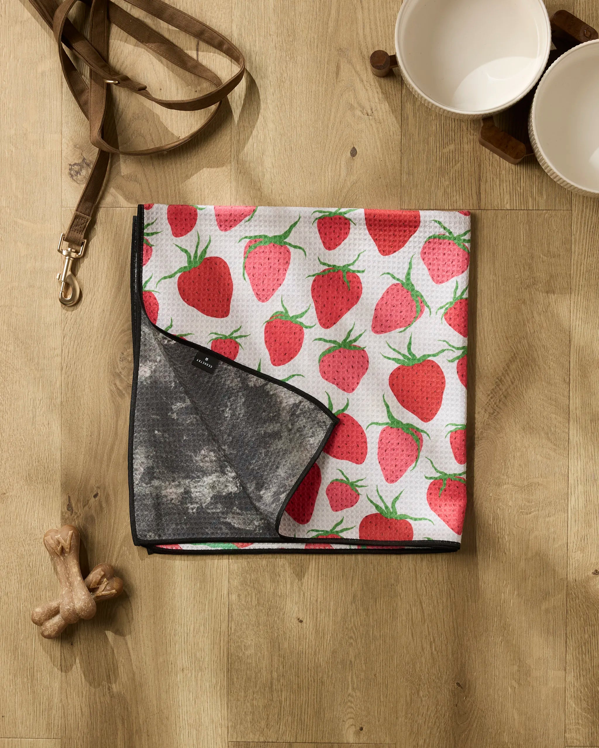 A dog leash, two white bowls, and two bone-shaped treats rest on a wooden floor beside the highly absorbent Geometry Strawberry Sniff dog towel with a strawberry pattern and dark lining.