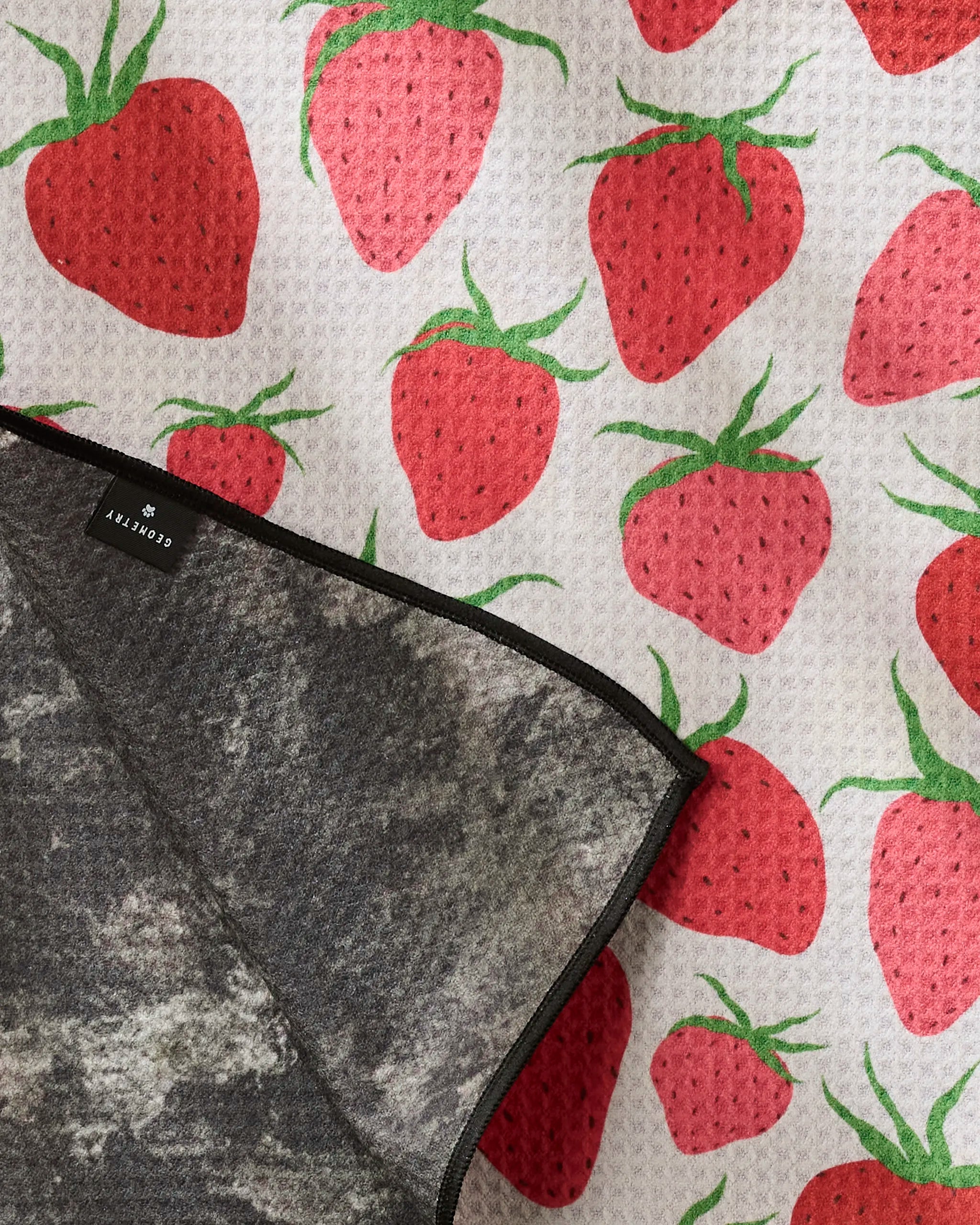 The Geometry Strawberry Sniff is a textured white dog towel with a red strawberry pattern, partially covered by a gray cloth with a black-edged COURTLY label, made from highly absorbent recycled materials.