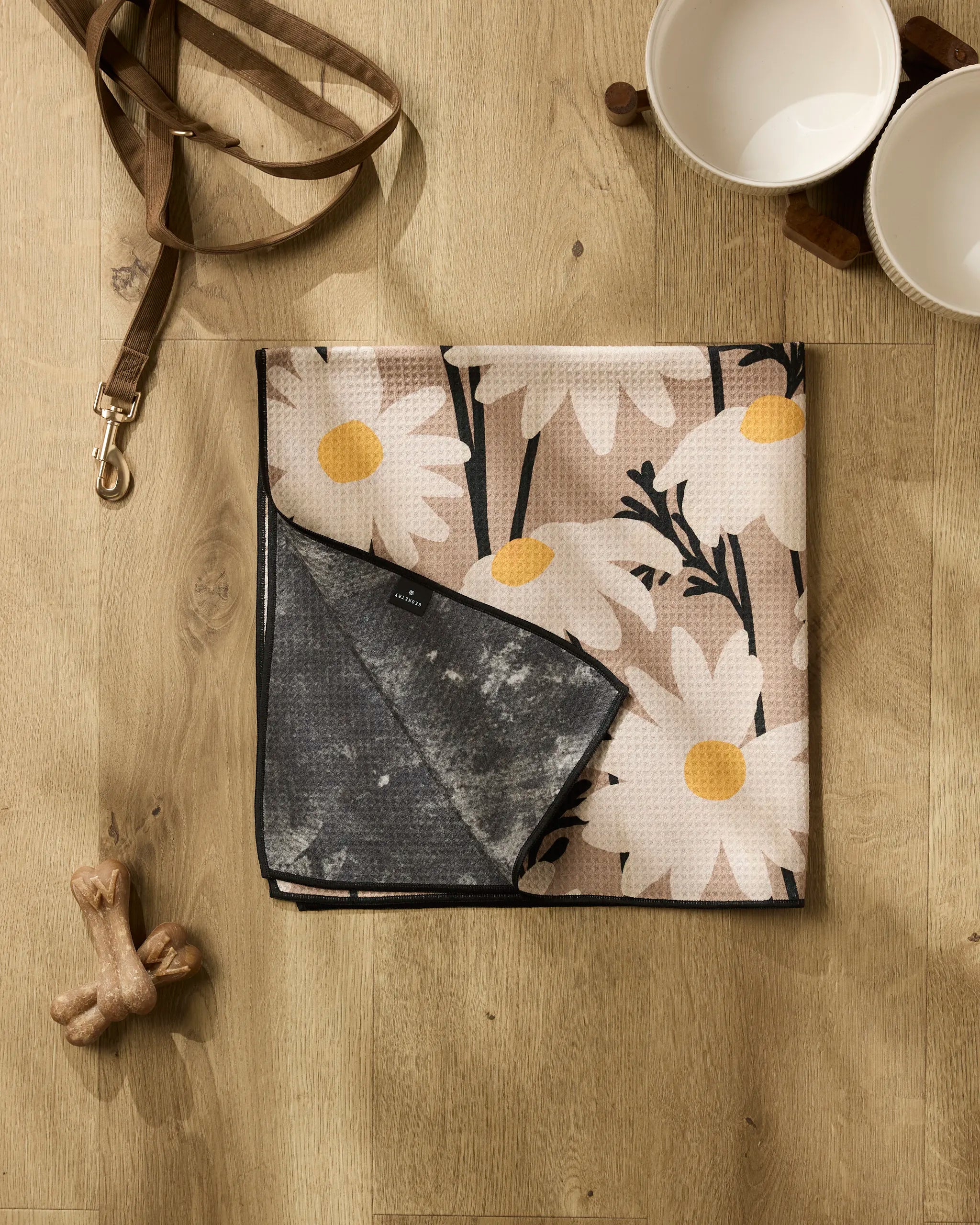 The Sweet Daisies dog towel by Geometry, featuring a floral design with white daisies, lies folded on a wooden floor beside a brown leash, ceramic bowls, and wooden dog bones.