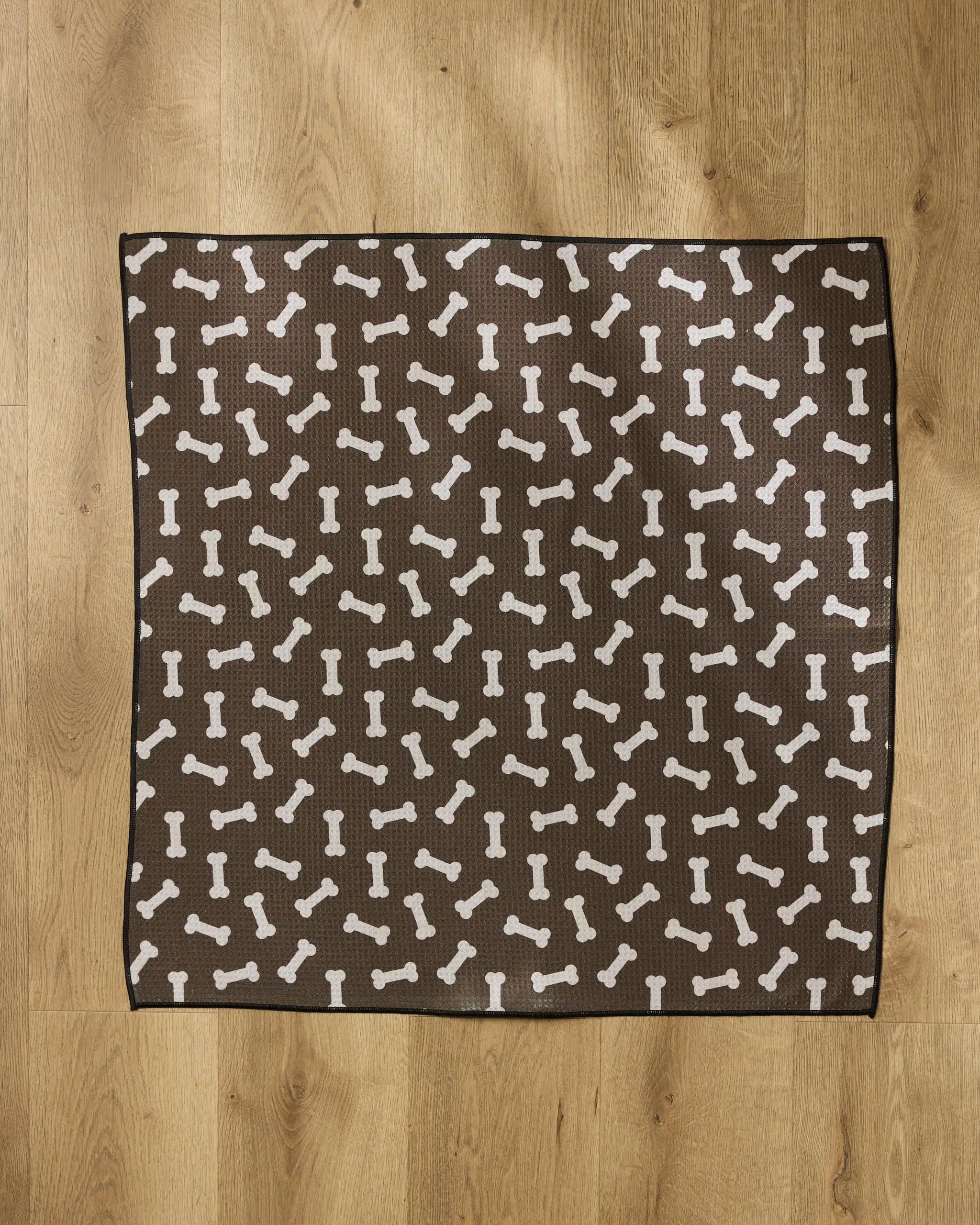 The Geometry Sweet Treats mat is a brown, square dog mat featuring a white bone pattern, made from absorbent towel material, and placed on a wooden floor.