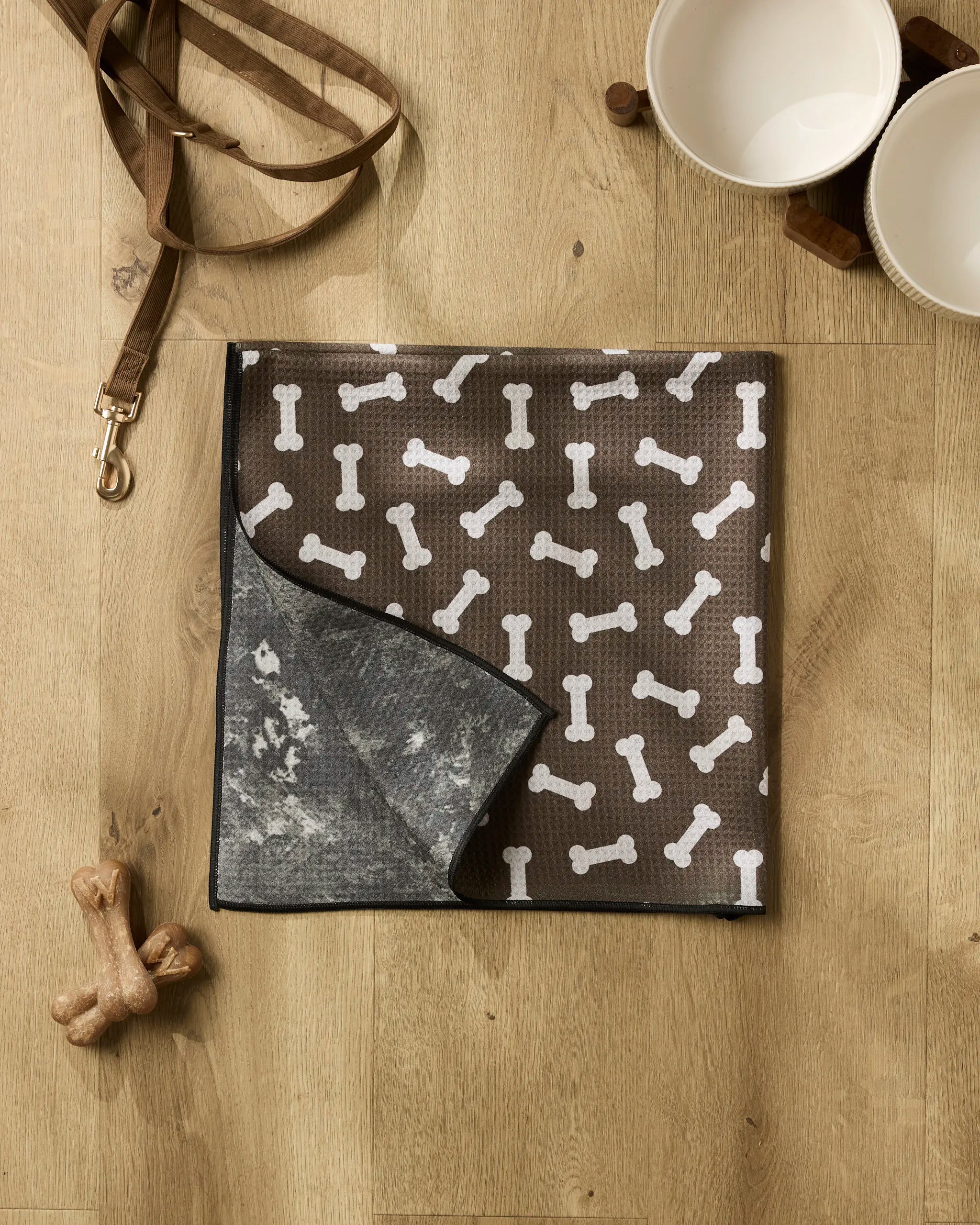 A brown dog mat with white bone patterns from Geometry's Sweet Treats collection lies on a wooden floor, joined by a recycled dog towel, leash, two ceramic bowls on a wooden stand, and two dog bone toys for a cozy corner.