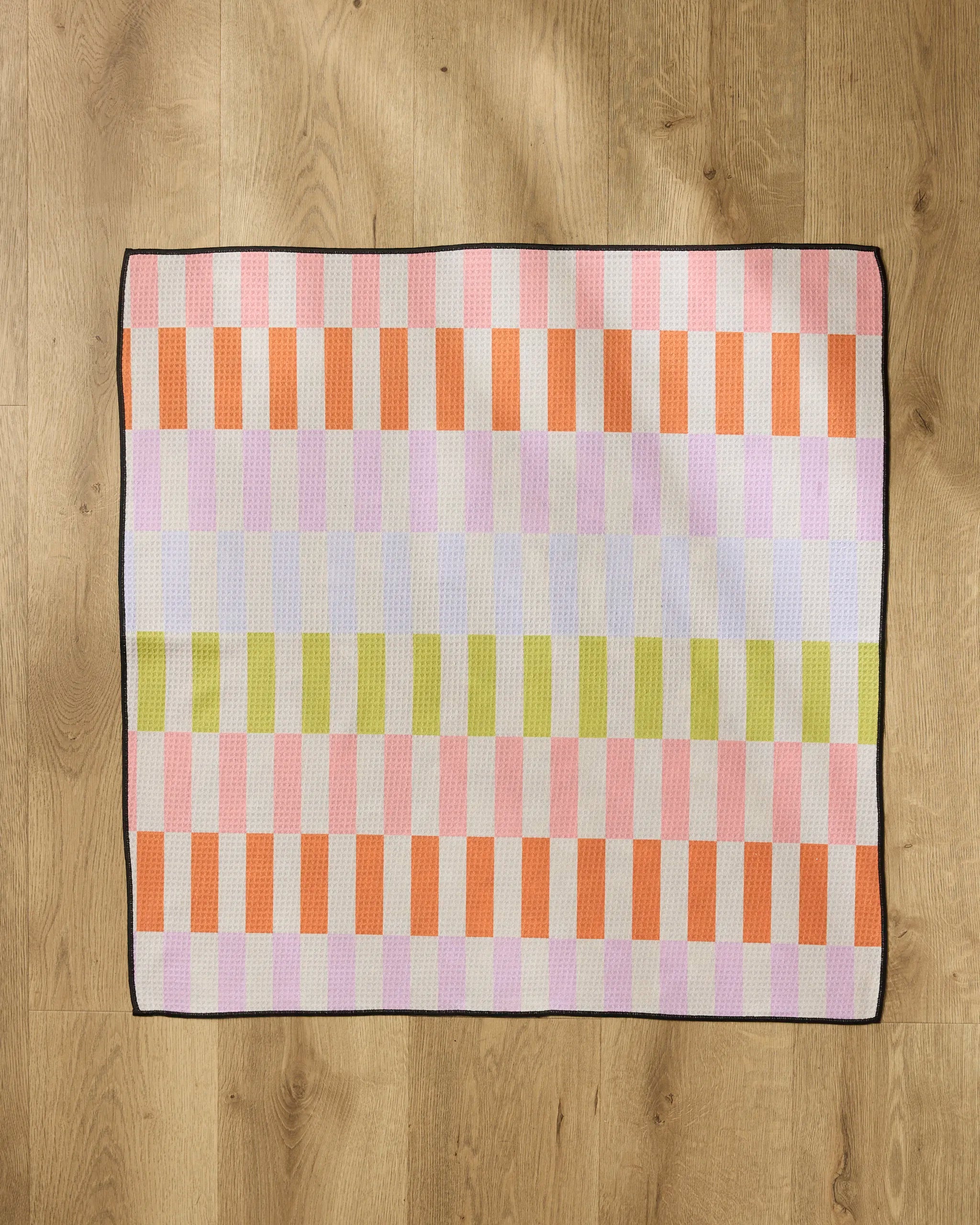 The Geometry Wag Wise mat features a checkered pattern in orange, pink, purple, and green stripes with a black border. Highly absorbent and rectangular, it adds a pop of color to any light wooden floor.