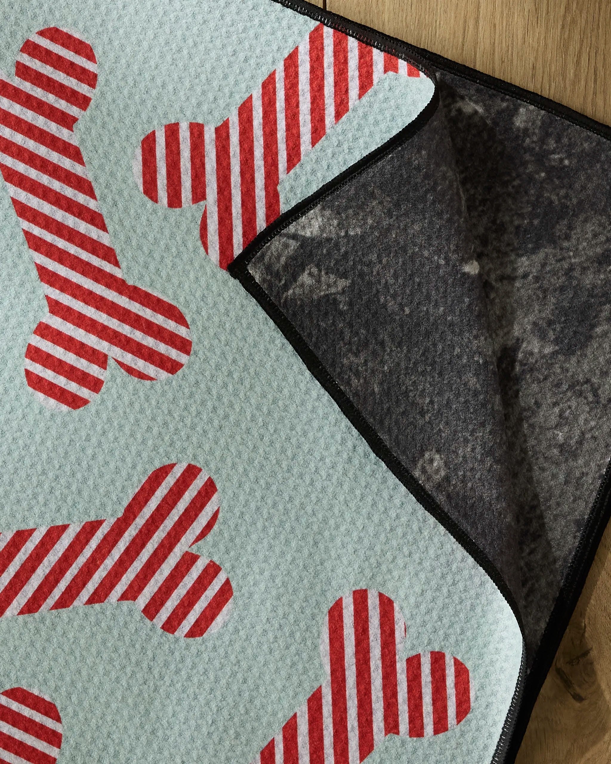 The Geometry Yuletide Pup pet mat features red and white striped bone patterns on light blue, and is shown partially flipped to display its dark grey, non-slip backing—crafted from recycled materials for an eco-friendly touch.