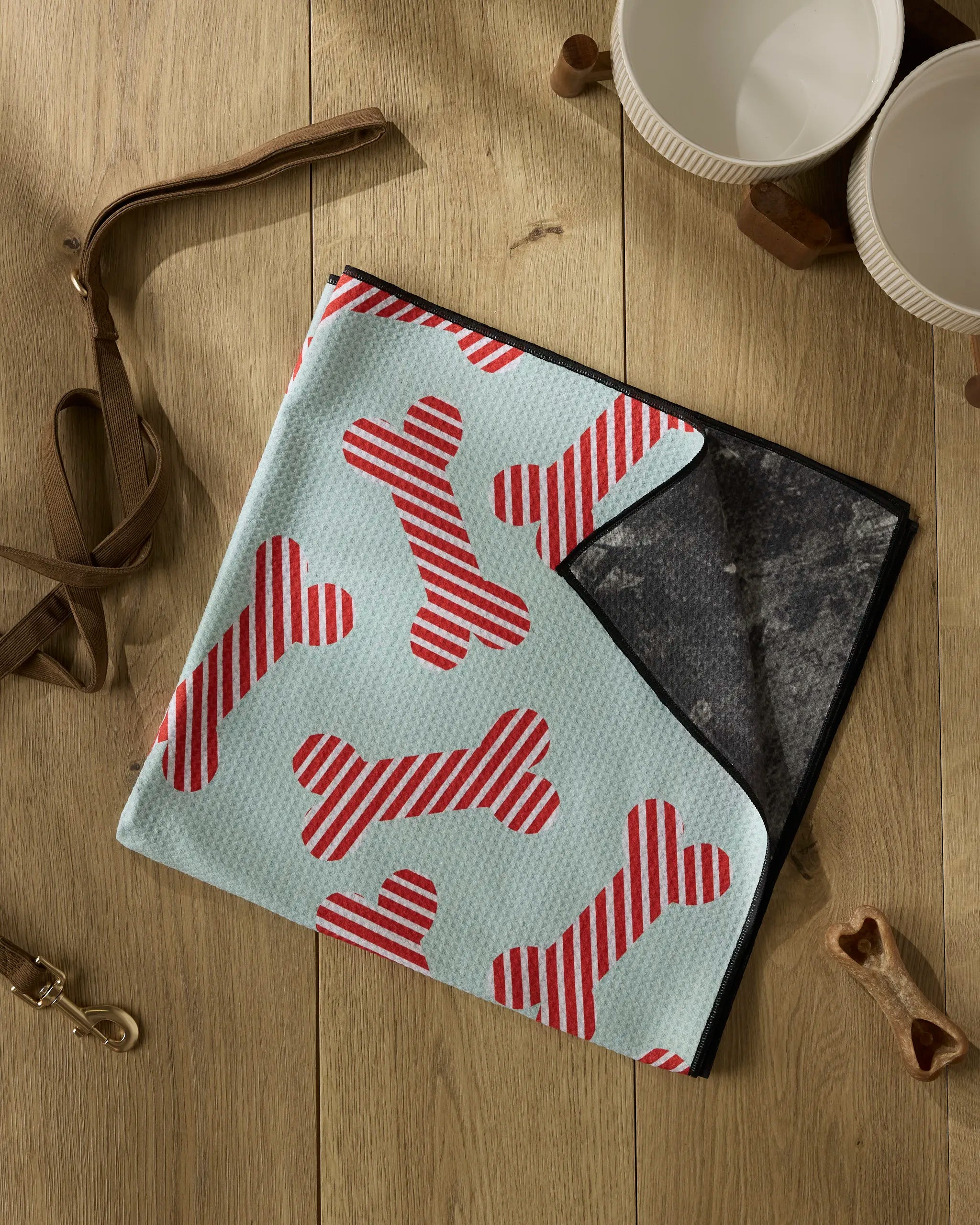 The Geometry Yuletide Pup, a light blue dog towel with red and white bone stripes made from recycled materials, is folded on wood beside two white bowls, a brown leather leash, and a gold dog collar clip.