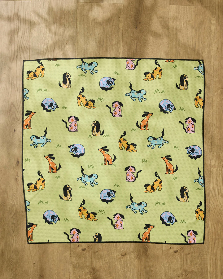 A Geometry "Summer Lawn" recycled dog towel with a black border is on a wooden floor. The green cloth features cartoon dogs in yellow, blue, and pink in various poses.