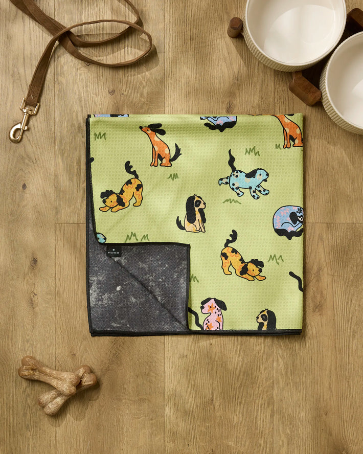 A Geometry "Summer Lawn" recycled green dog towel with colorful cartoon dogs is folded on a wooden floor alongside a leather leash, two dog bowls, and two bone-shaped chews.