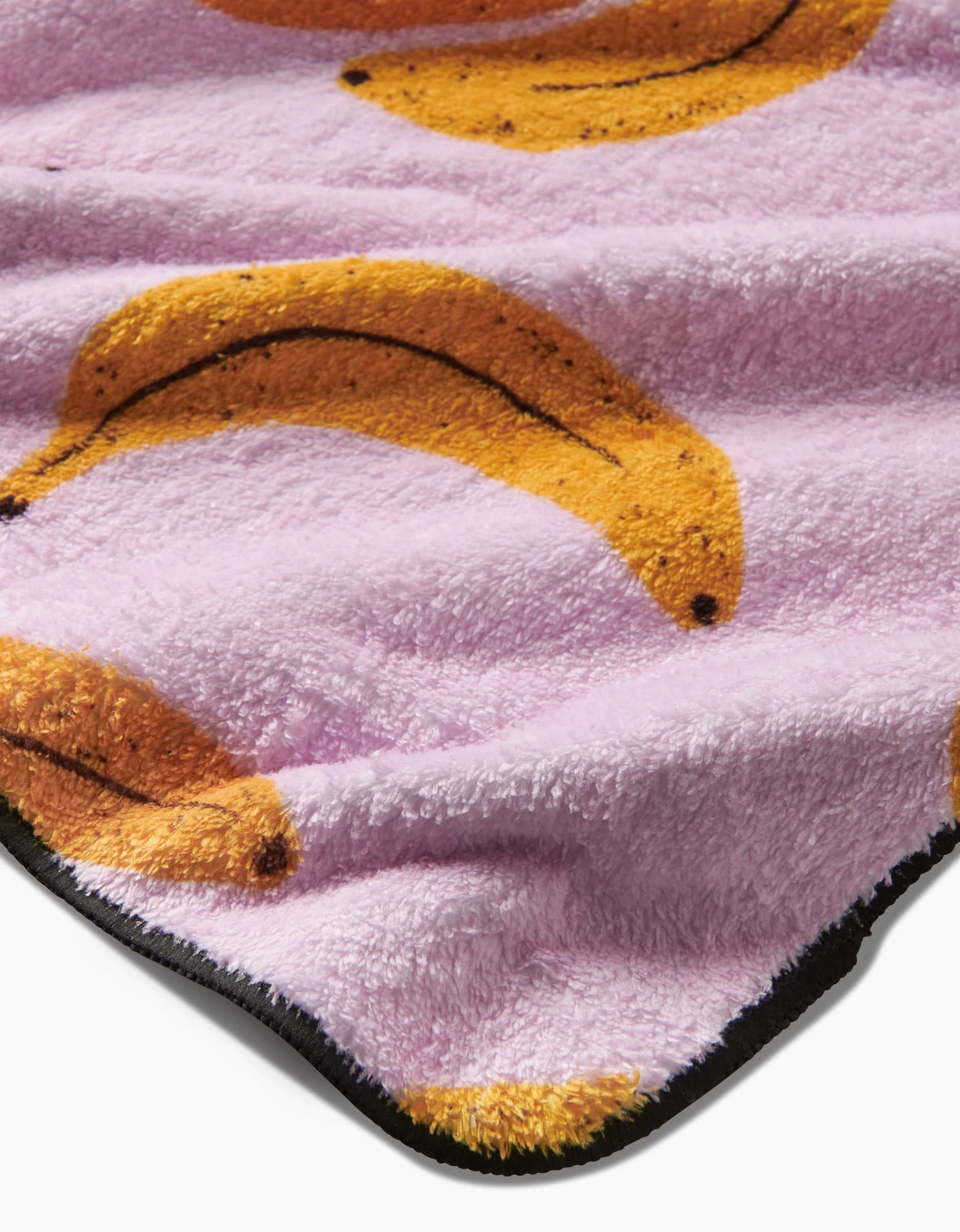 The Geometry Pupnana blanket features soft, fluffy fabric made from recycled materials, a purple background with yellow banana patterns, and a stylish black edge trim.