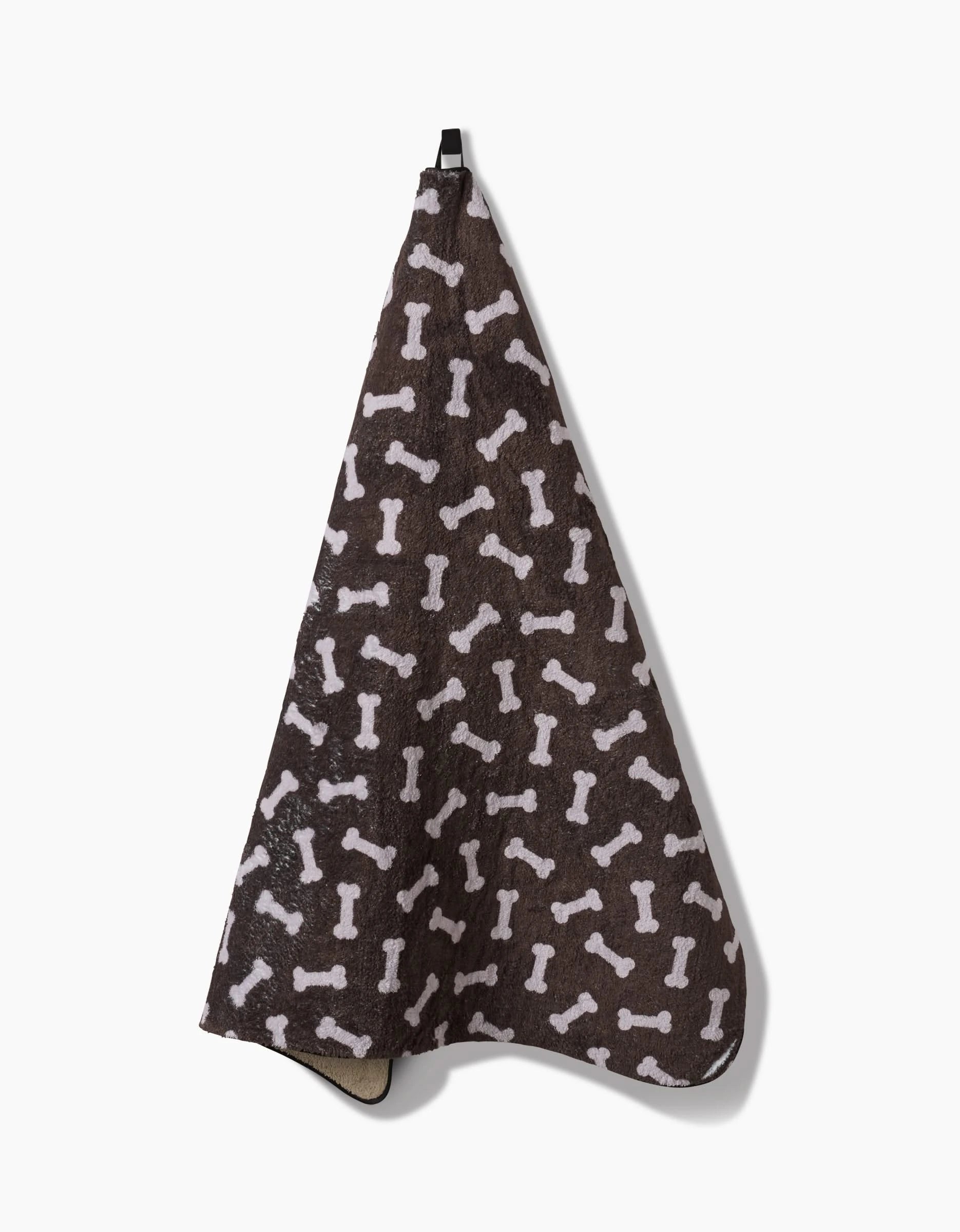 The Sweet Treats towel by Geometry is a plush, ultra-absorbent dog towel in dark brown with white bone patterns, made from recycled materials. It hangs from a black loop and is shown against a plain white background.