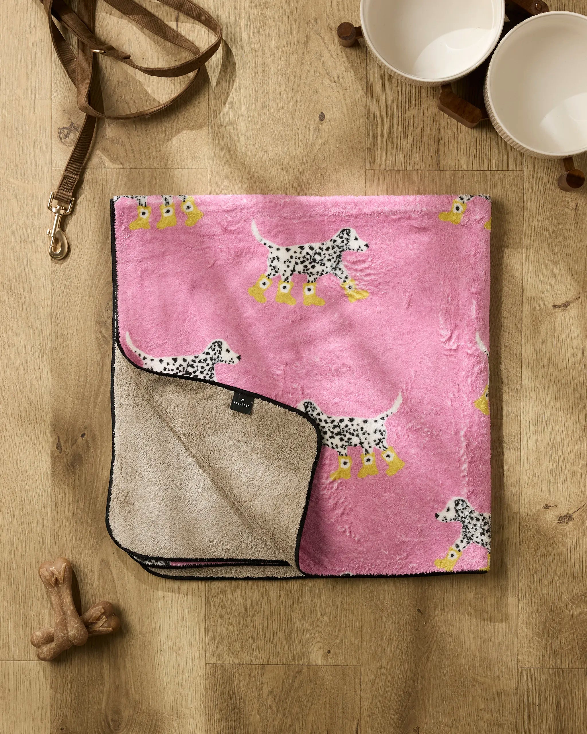The Geometry Dalmatian In Boots Pink dog towel with a Dalmatian pattern is laid out on a wooden floor next to a leather leash, two white bowls, a dog bone toy, and a dog-shaped treat.