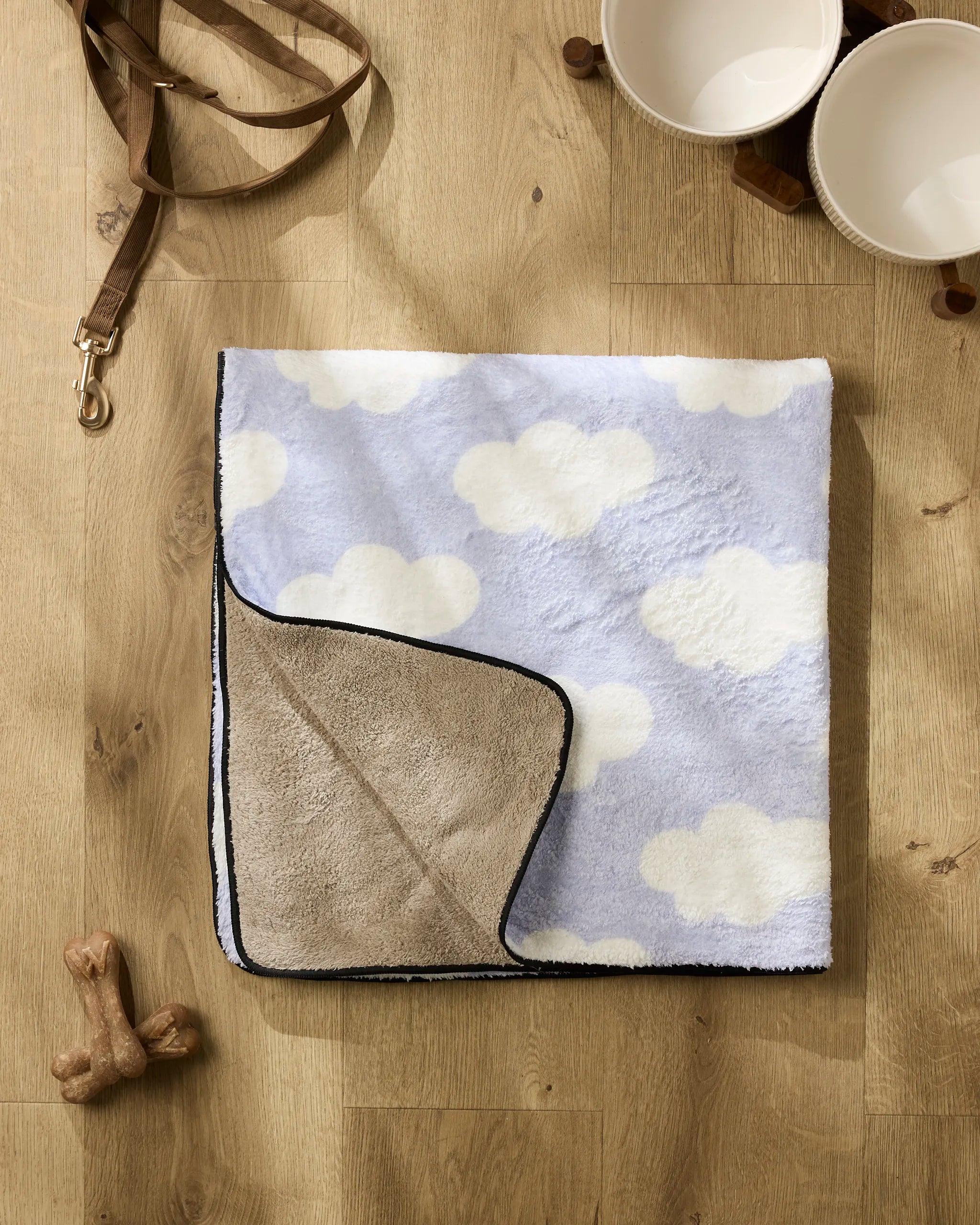 The Dreamy Clouds plush dog towel by Letters By Shells, featuring cloud patterns, is folded on a wooden floor next to a brown leash, two white pet bowls, and two dog bones.