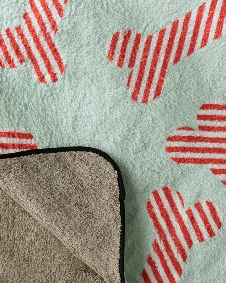 The Geometry Yuletide Pup is a plush dog towel with red and white striped bone patterns on a light background, and features one corner folded over to show its solid gray underside.