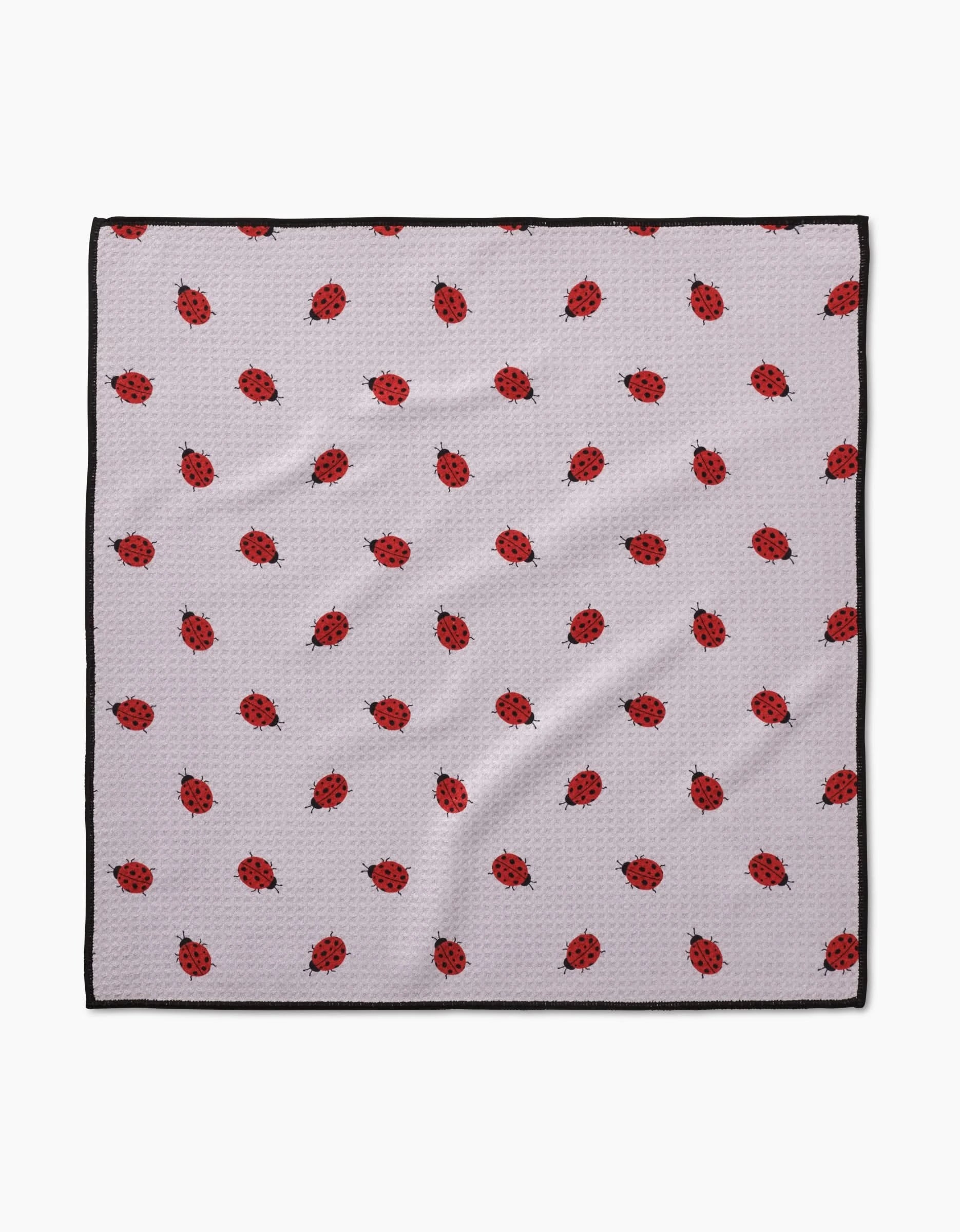 The Geometry Bugsy Paw Towel features a white background with red ladybugs, bordered by a thin black edge. Made from ultra-absorbent Geoweave fabric, the towel is slightly textured and wrinkled in appearance.