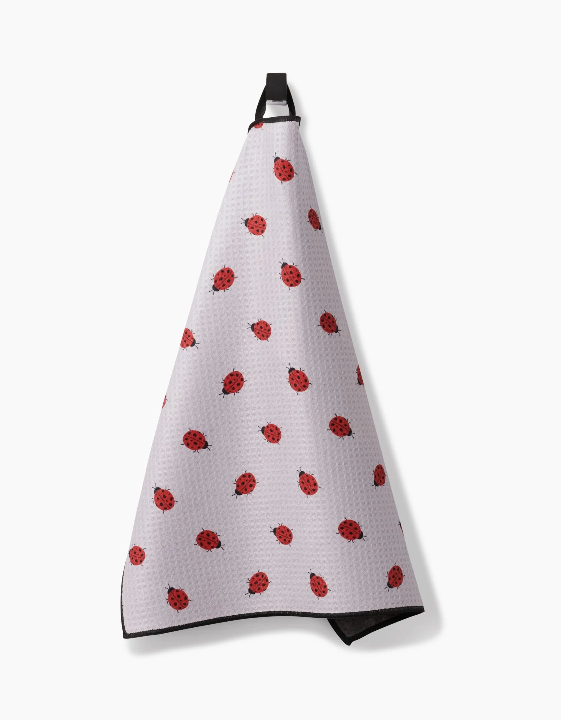 The Geometry Bugsy kitchen towel, featuring a white ultra-absorbent fabric with red ladybug motifs, hangs from a black loop against a white background.