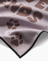 A close-up of the Geometry Fur Ever Friends Paw Towel shows its light purple Geoweave fabric with brown paw prints, bold FRIENDS text, and a sleek black border for ultra-absorbency.