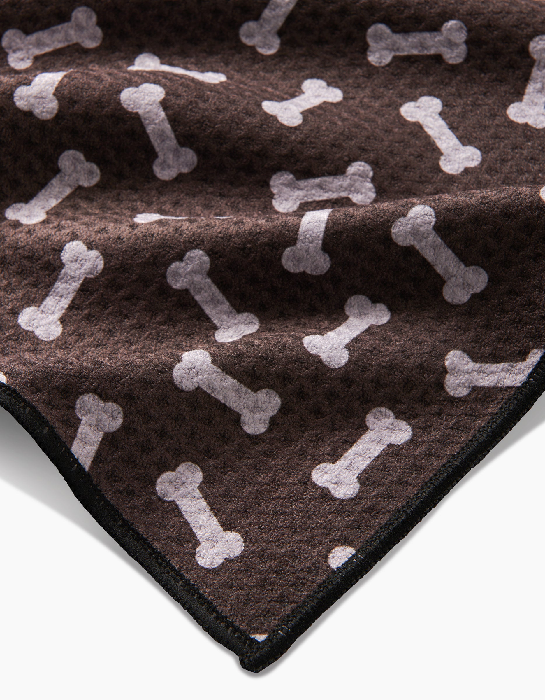 The Geometry Sweet Treats is a soft, brown fleece blanket made from recycled materials, featuring a repeating white dog bone pattern and black stitched edge, shown partially folded.