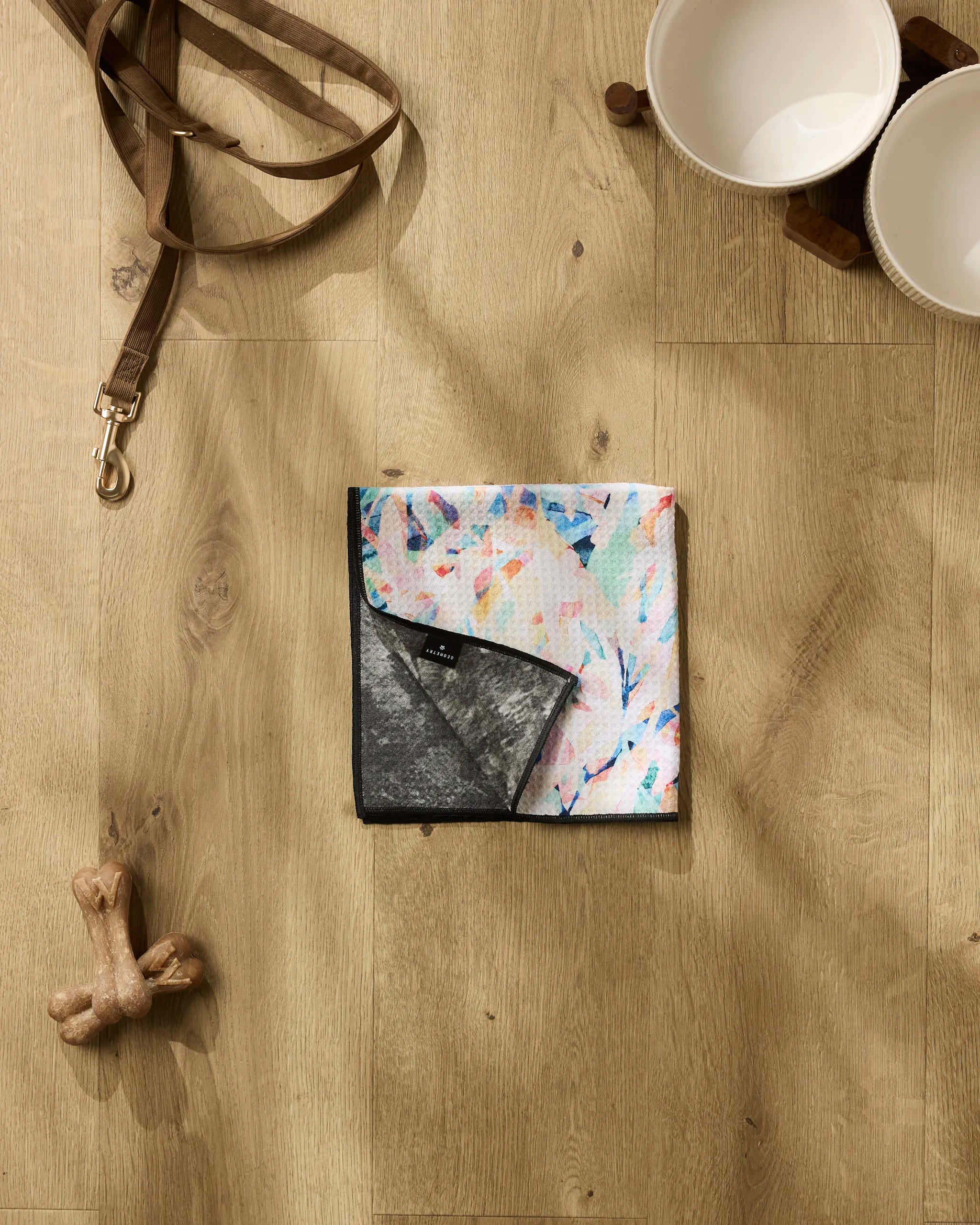 A flat lay of pet accessories on wood floor: a brown leash, two white bowls on a stand, dog bone toy, and the Abstract Jungle bandana by Rachel Parker—colorful with black underside—plus an ultra-absorbent towel for quick paw cleanup.