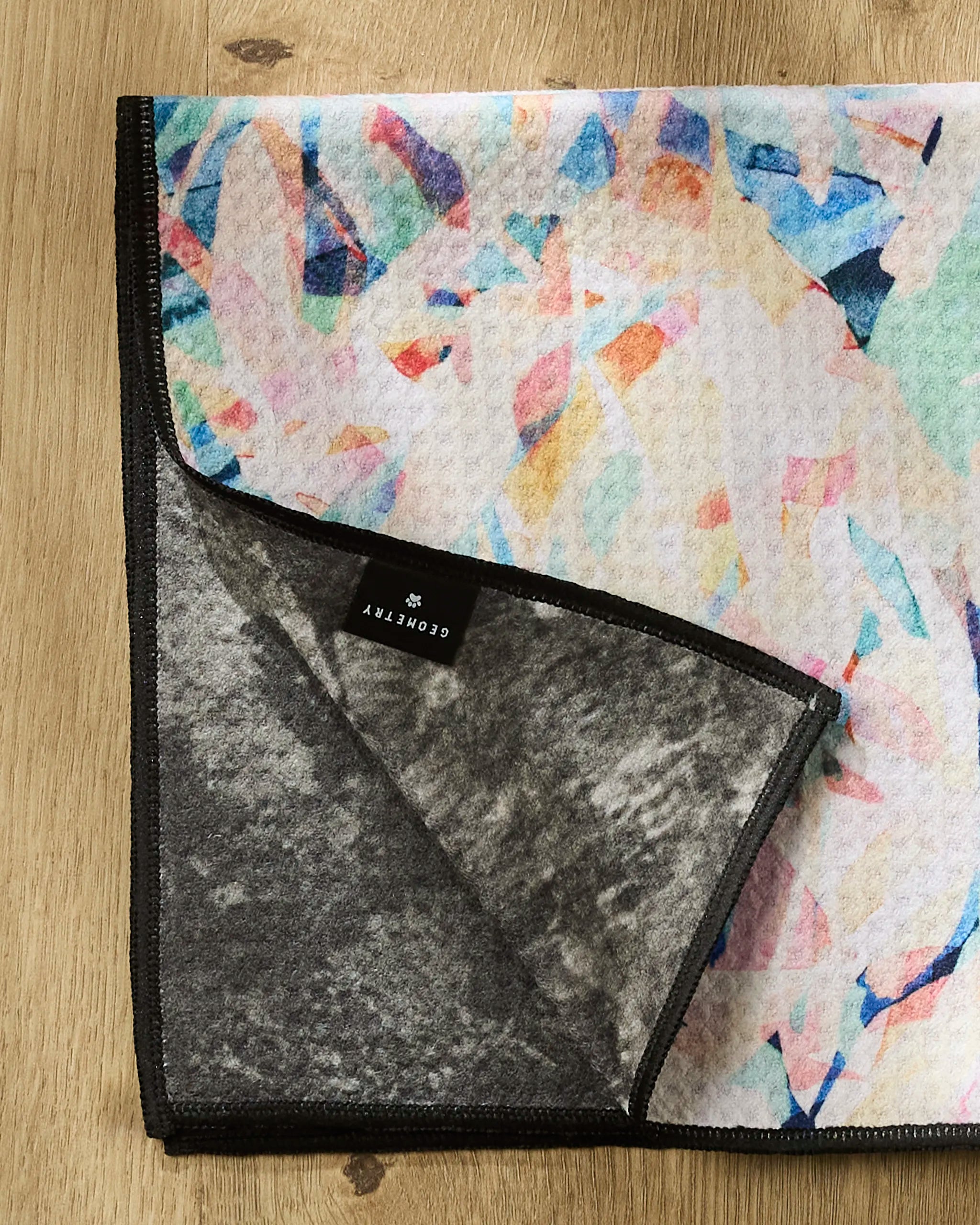 The Rachel Parker "Abstract Jungle" dog towel features a colorful abstract pattern with black edges, a gray underside, and a black GEOMETRY tag, shown folded at one corner on a wooden surface.