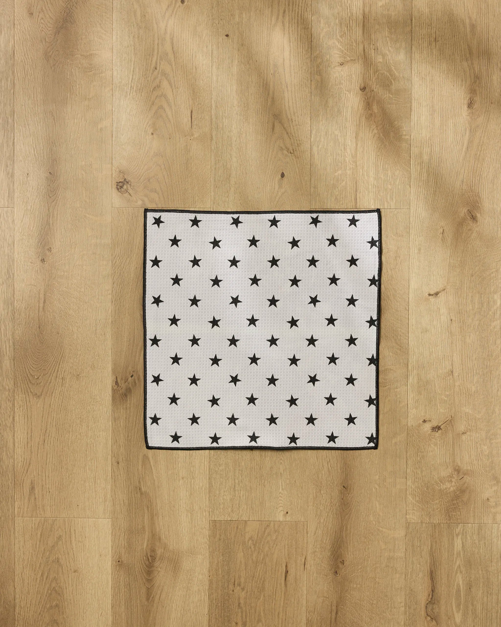 The Geometry Bark towel, ultra-absorbent and eco-friendly, features a white square design with evenly spaced black stars and black borders—perfect for pets. Made from recycled materials and pictured here on a light wood floor.