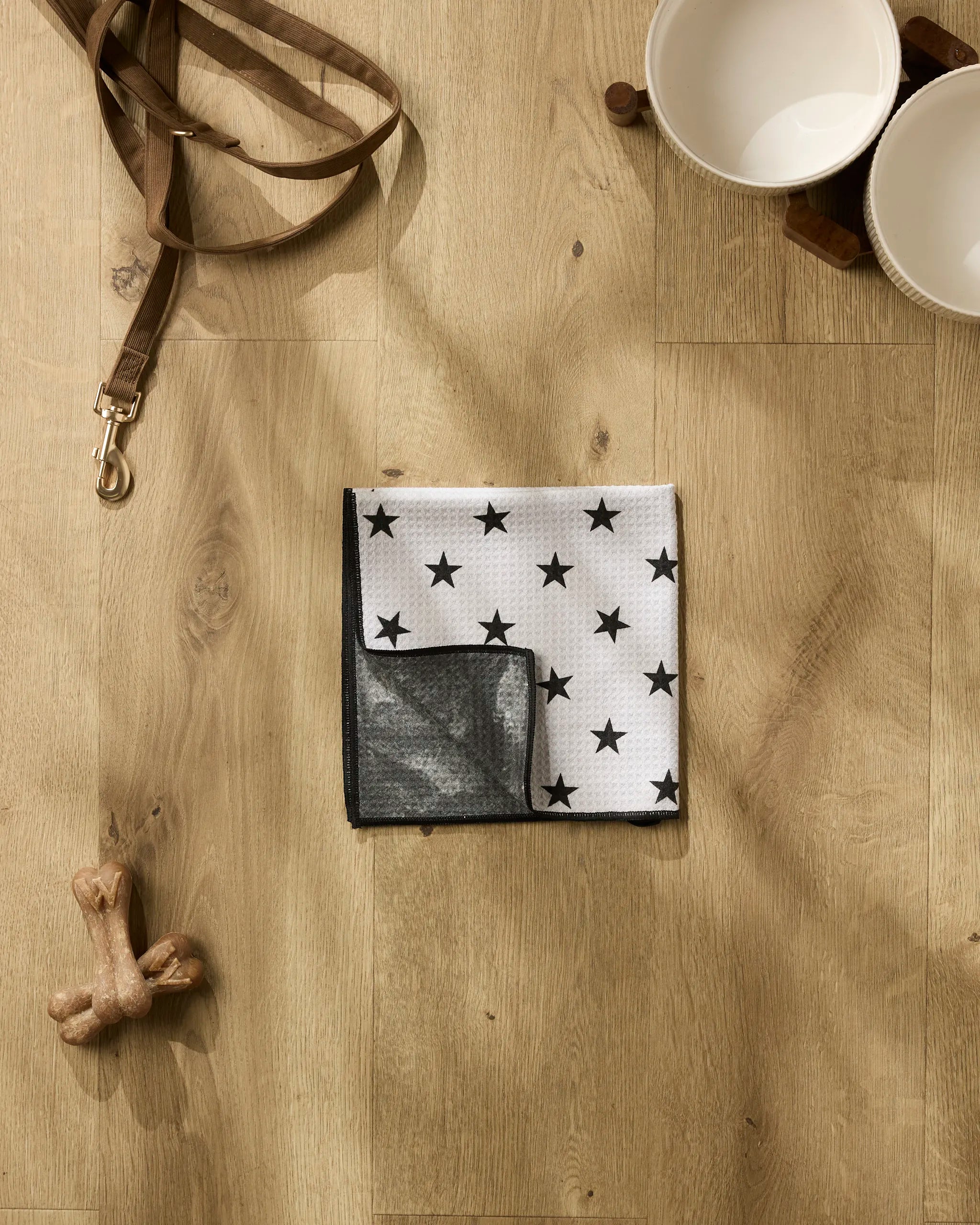 The Bark collection by Geometry features dog accessories on a wooden floor: a brown leash, two white bowls with a wooden stand, a bone-shaped chew toy, and an ultra-absorbent paw towel with black stars.