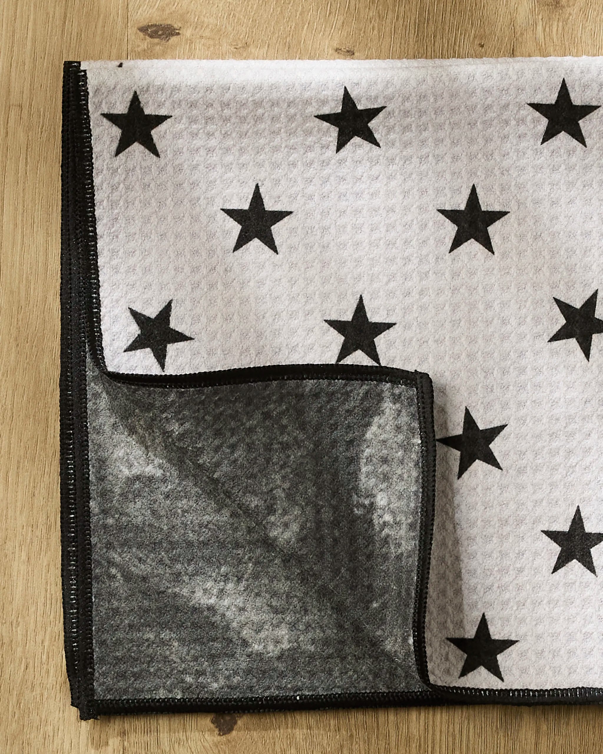 The Geometry Bark towel, made from recycled materials, features a white textured surface with a black star pattern, black trim, and a gray underside. It’s shown folded on a wooden surface.