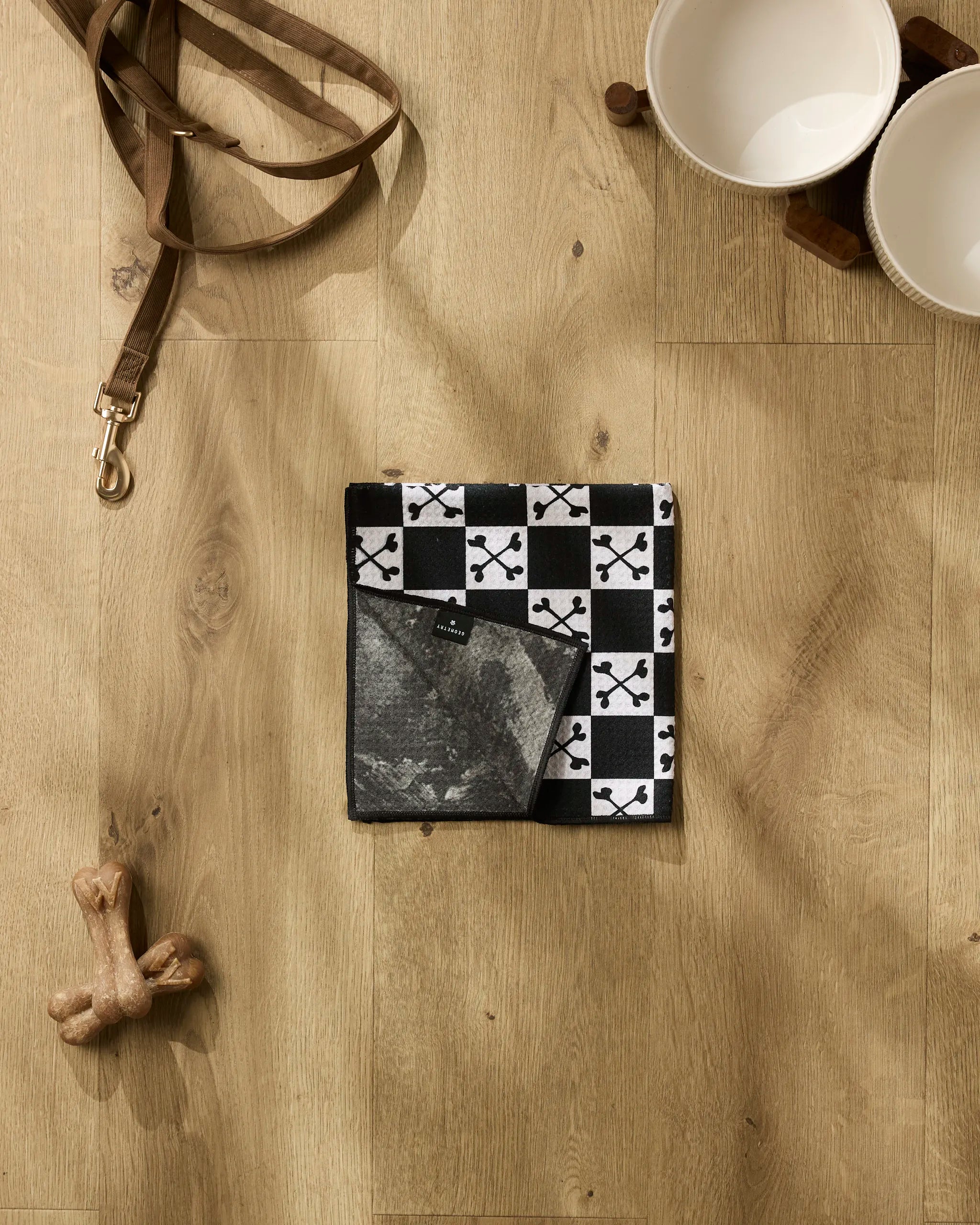 The Charlie Rowan Designs Boneyard White Checkerboard towel, crafted from ultra-absorbent recycled materials, rests folded on a wooden floor beside a brown leash, two ceramic dog bowls, and a dog bone toy.