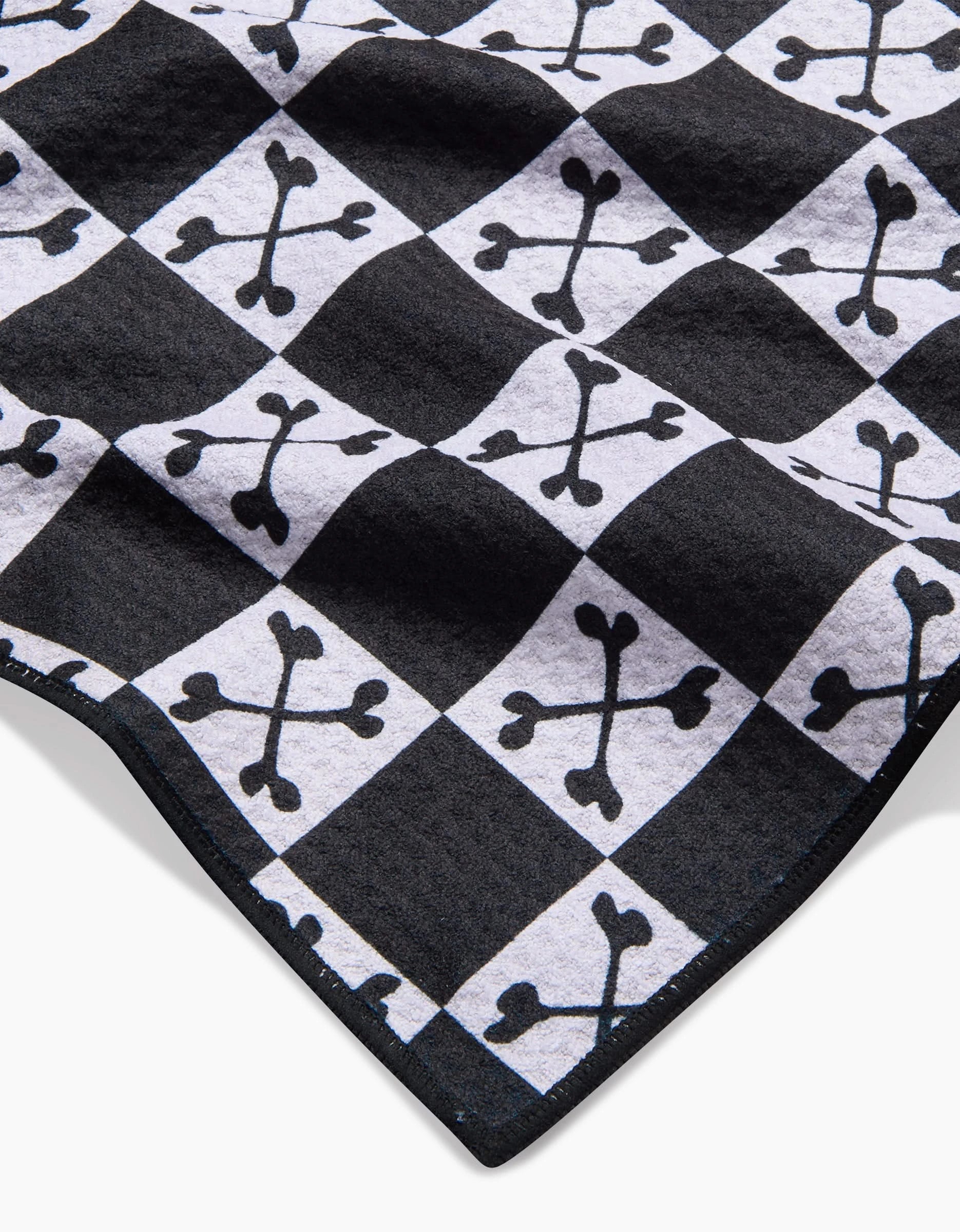 A close-up of Charlie Rowan Designs Boneyard White Checkerboard fabric shows its black and white checkered pattern with crossbones featuring heart-shaped ends, all on an ultra-absorbent towel made from recycled materials.