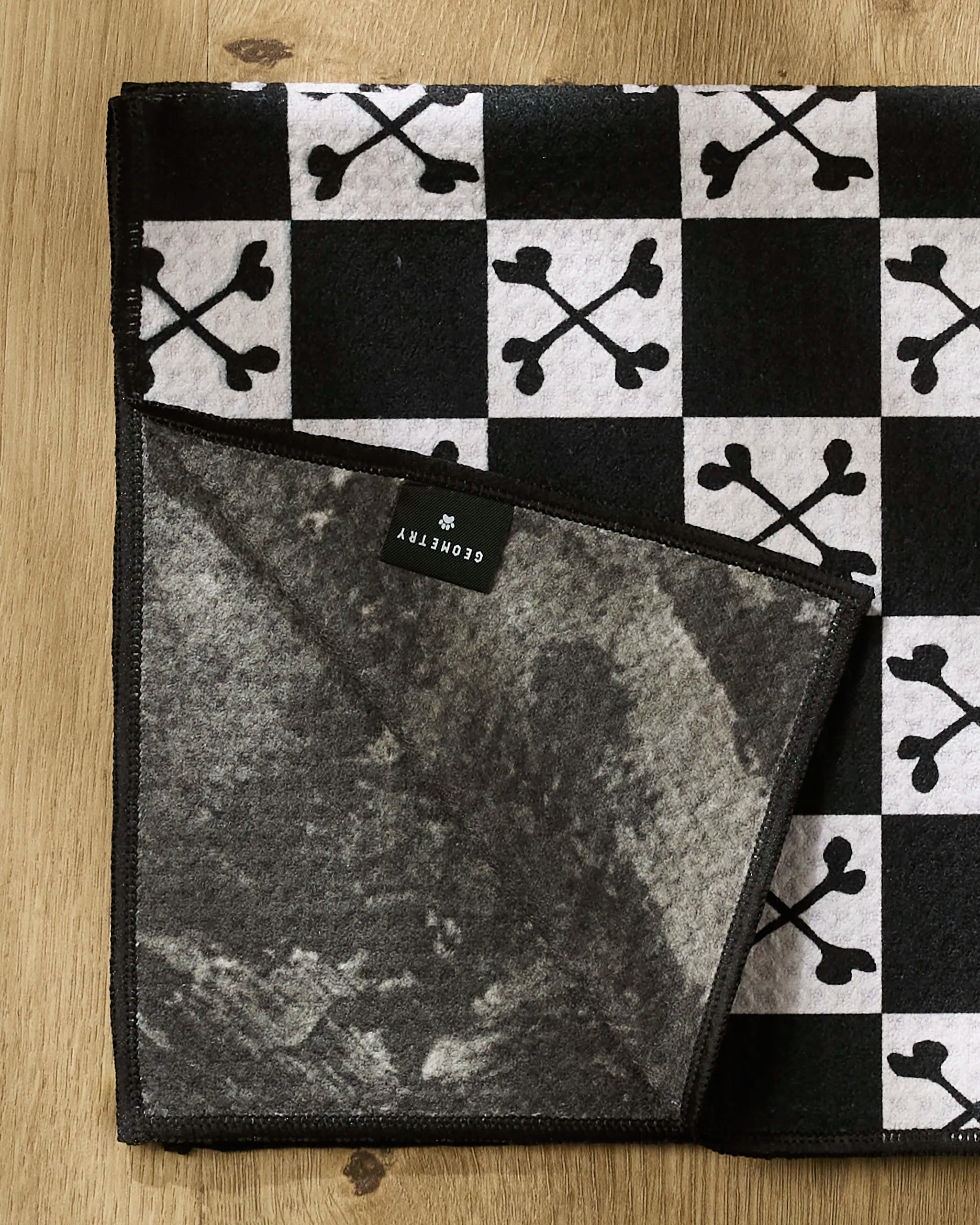 The Boneyard White Checkerboard blanket by Charlie Rowan Designs features a black and white checkered skull pattern, folded on wood. Made from recycled materials, its flipped corner shows a gray marbled underside with a black COVENTRY tag.