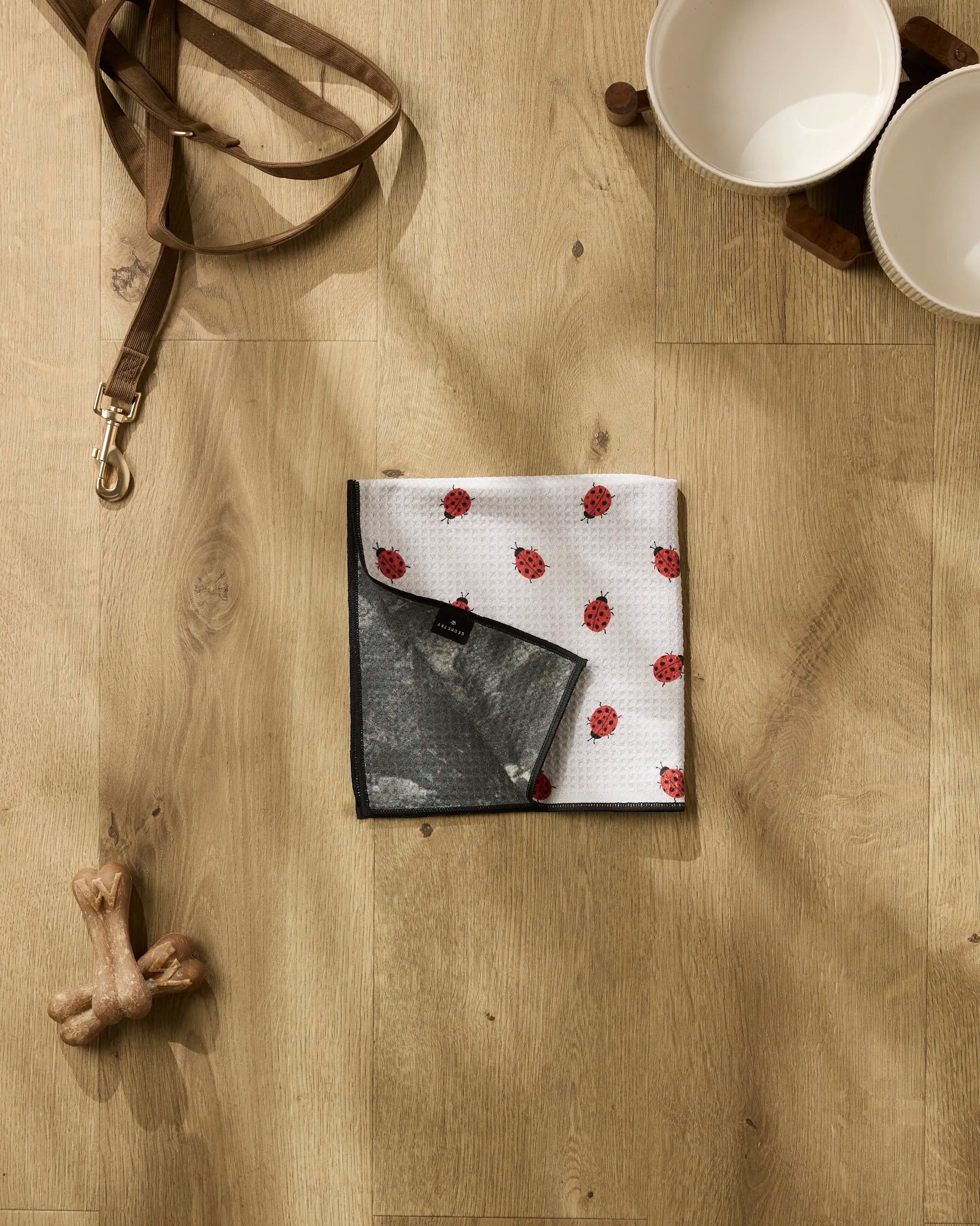 Geometry’s Bugsy ultra-absorbent dog towel with red ladybug prints is displayed on a light wooden floor, alongside a leash, a dog bone toy, and two white bowls on a wooden stand.