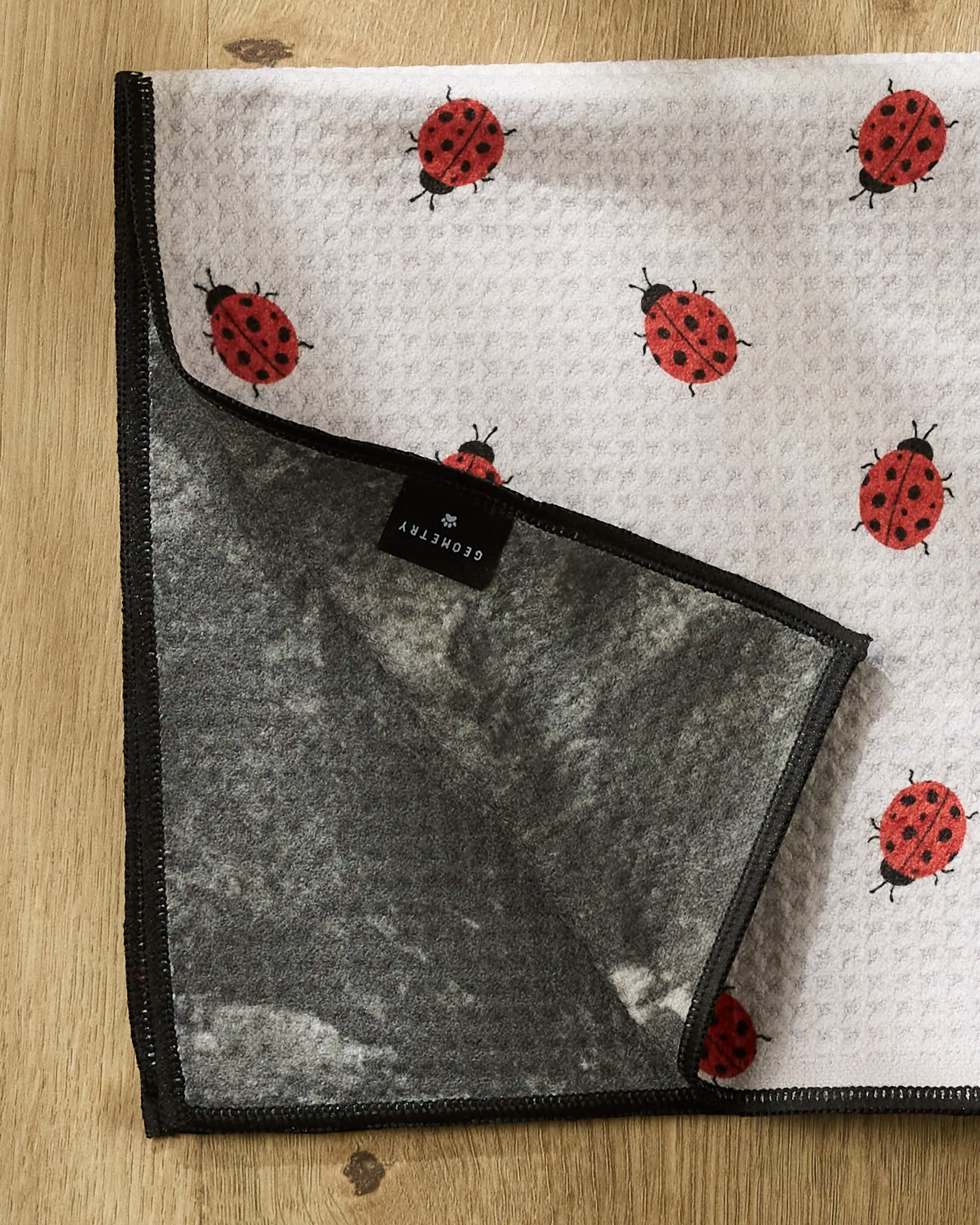 The Geometry Bugsy paw towel features a white surface with red ladybug patterns and a gray underside, folded at one corner. Ultra-absorbent and stylish, it's perfect for your pet’s next bath or adventure.