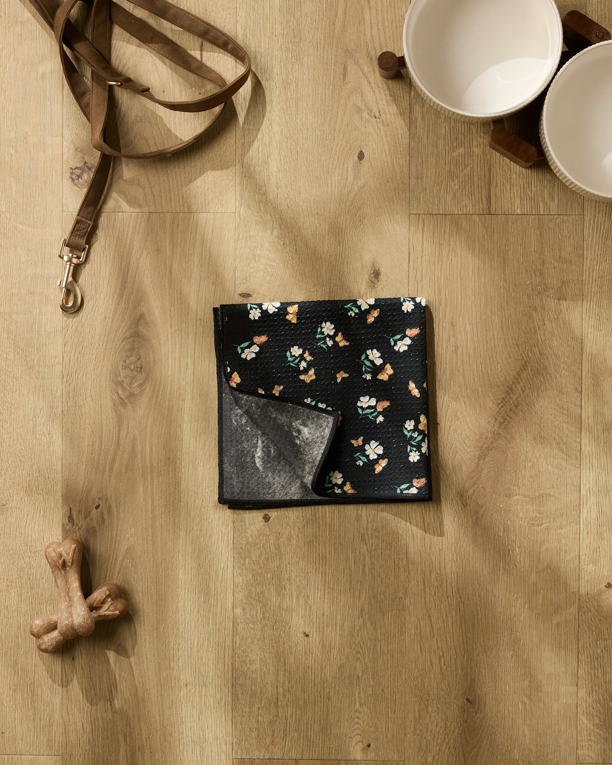 Danelys Sidron's "Butterfly Garden" black floral pet bandana, tan leash, white dog bowls on a wooden feeder, two bone toys, and an ultra-absorbent dog towel are arranged on a light wood floor.