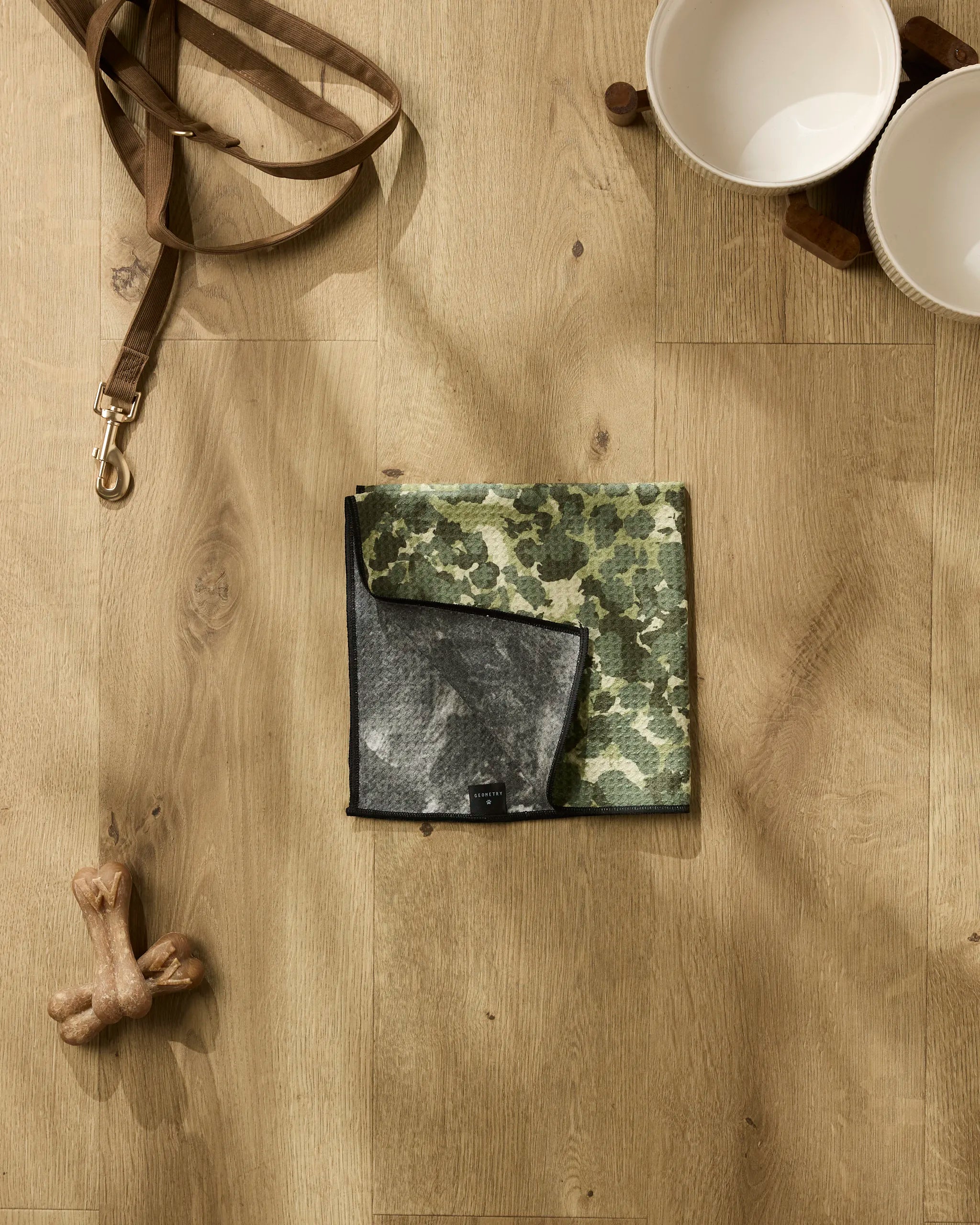 A Geometry "Camo" dog mat with a black edge is centered beside an ultra-absorbent dog towel, surrounded by a leash, two white bowls, and a bone-shaped toy on a light wood floor.