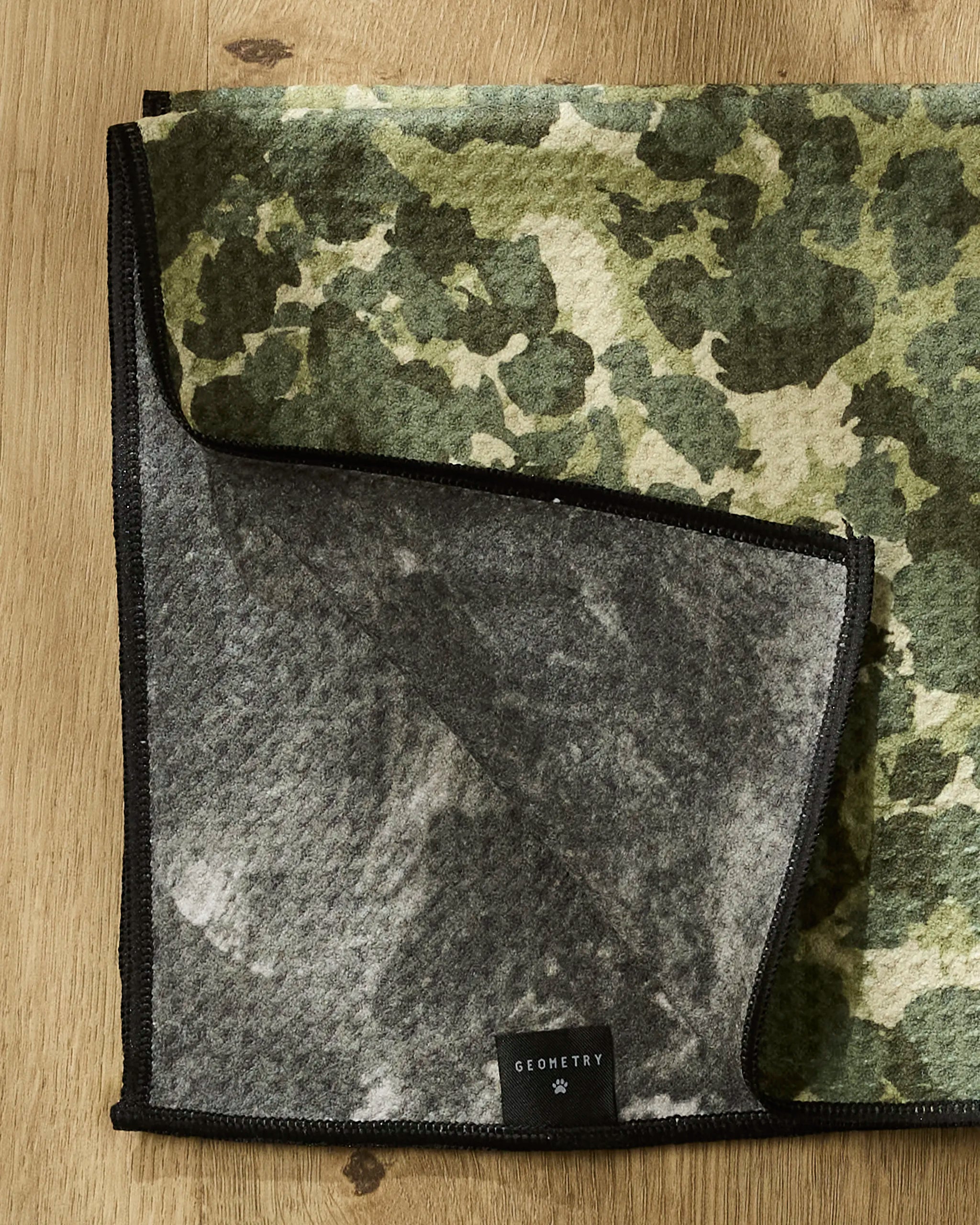 A green and beige camo-patterned ultra-absorbent dog towel from Geometry, named "Camo," lies on wood with a flipped corner showing a gray underside and a black GEOMETRY label.