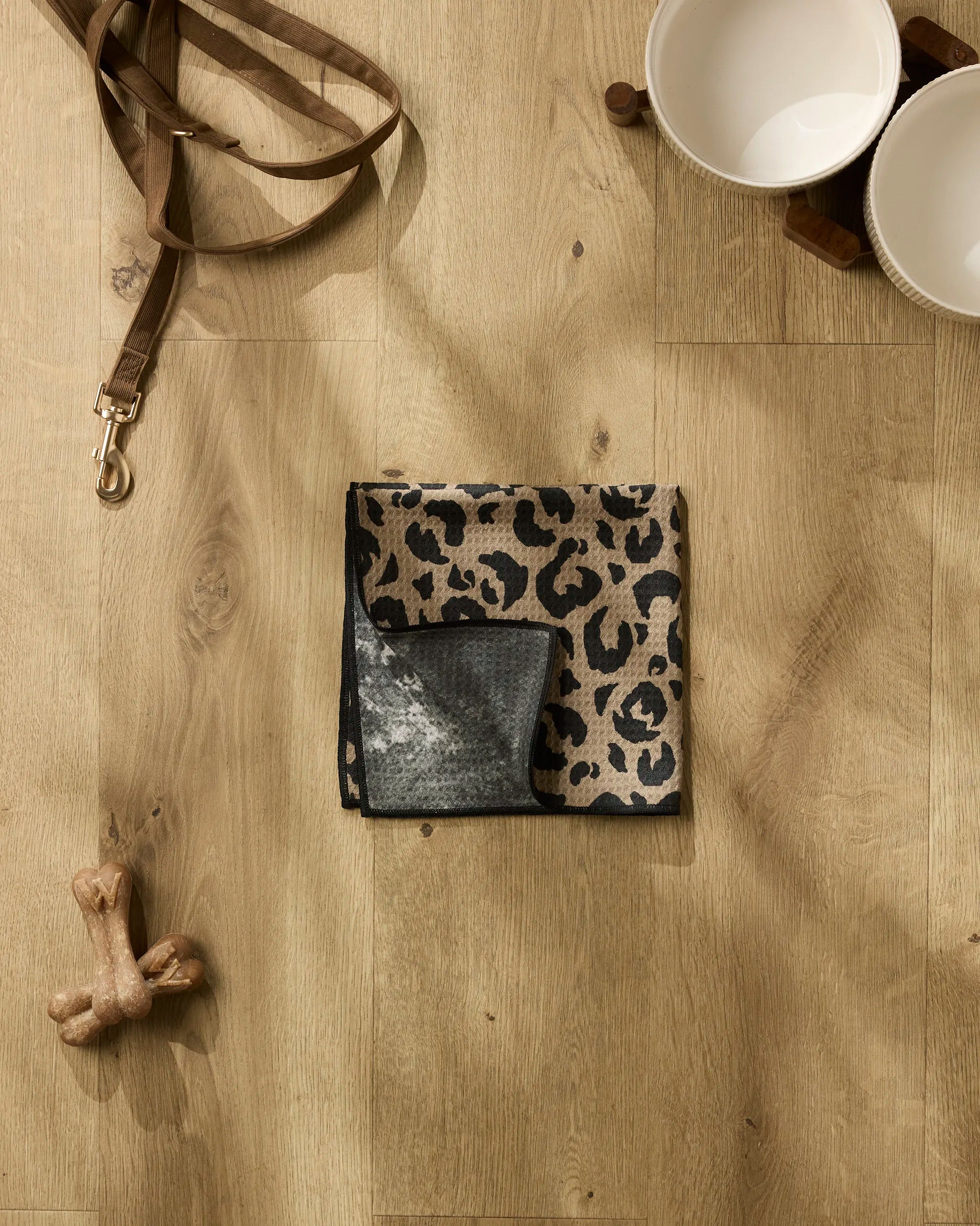 A flat lay of the Geometry Cheetah Hound leash, two white bowls on a wooden stand, an ultra-absorbent dog towel, leopard print cloth, and bone-shaped treats displayed on a light wood floor.