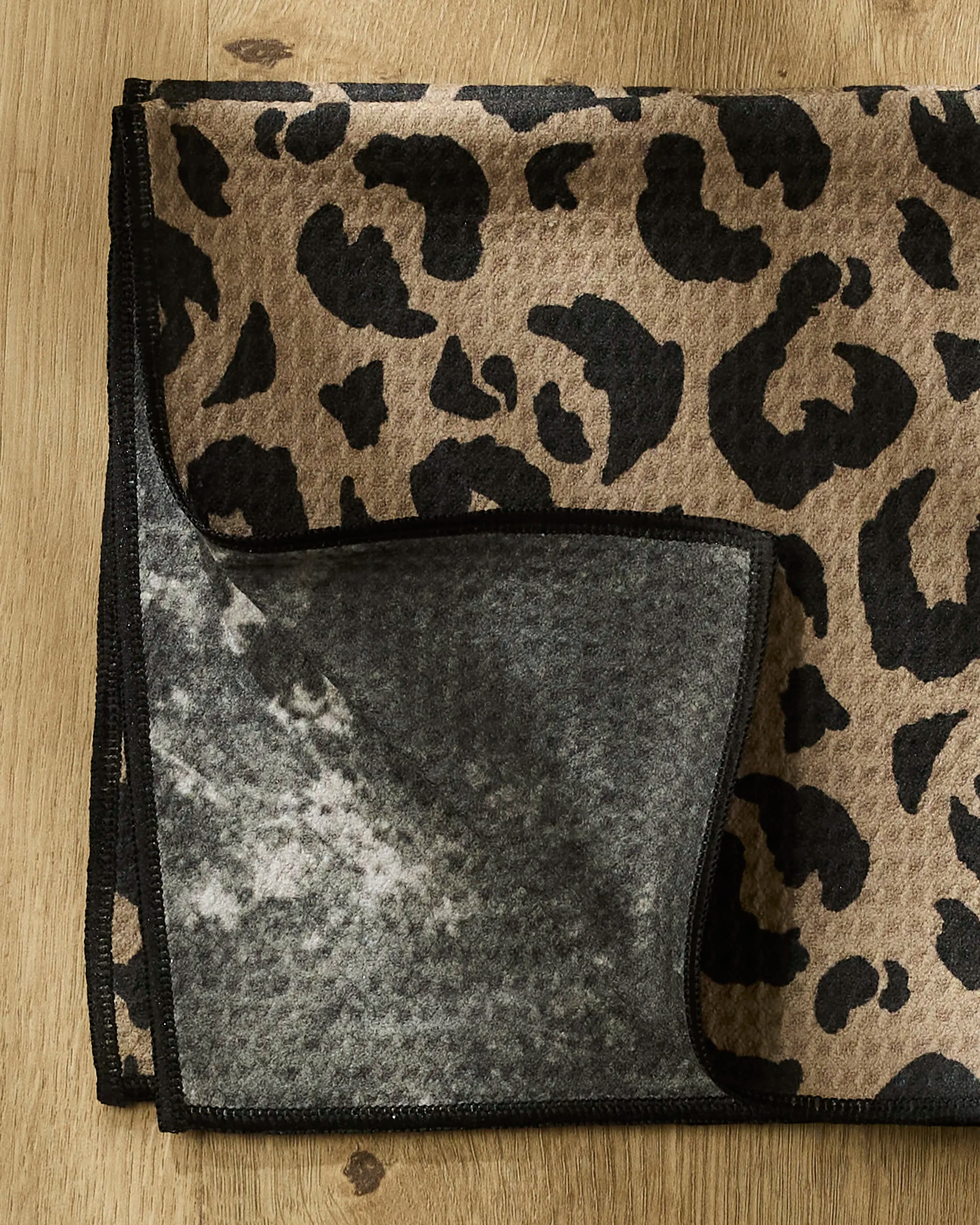 A Geometry Cheetah Hound Paw Towel, featuring a brown and black cheetah print on one side and plain gray on the other, is folded on a wooden surface with both sides visible.