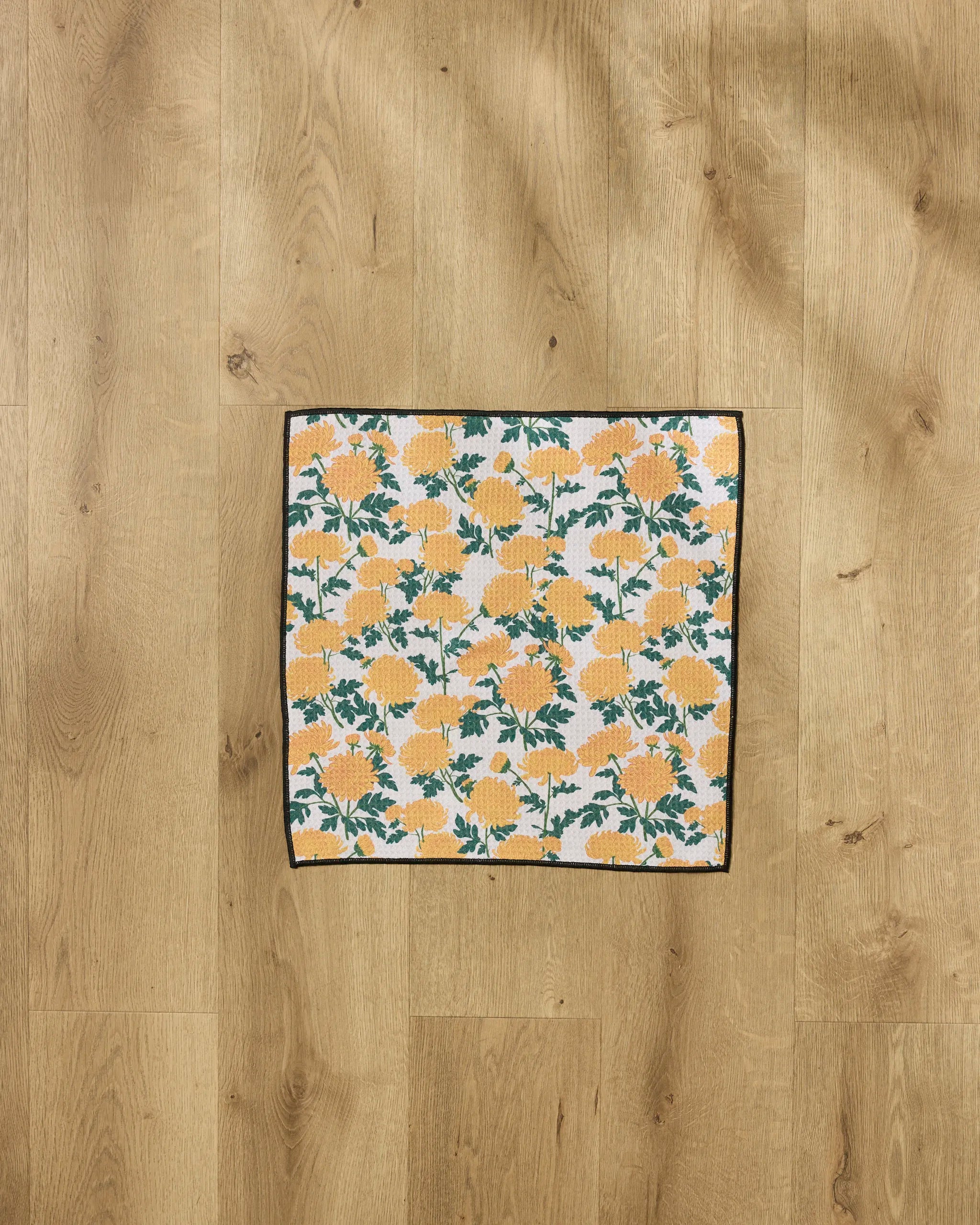 The Chrysanthemum towel by Vaishali Design Studio features ultra-absorbent rectangular cloth with yellow floral and green leaf patterns on white, shown flat on a light wood floor — a stylish Paw towel.