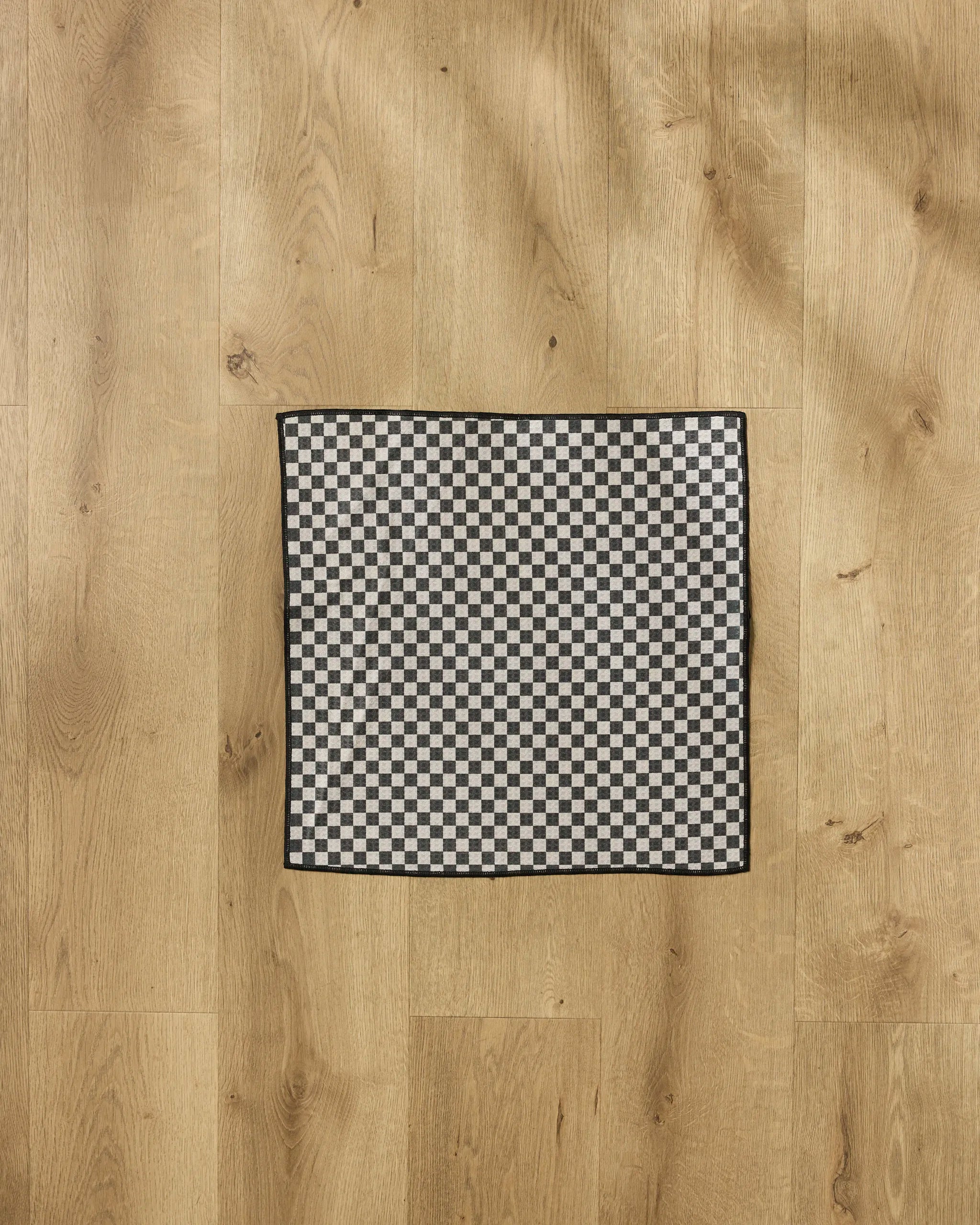 The Geometry Cool Pup towel, featuring a black and white checkered design made from recycled PET, is neatly folded on a light wooden floor and viewed from above.