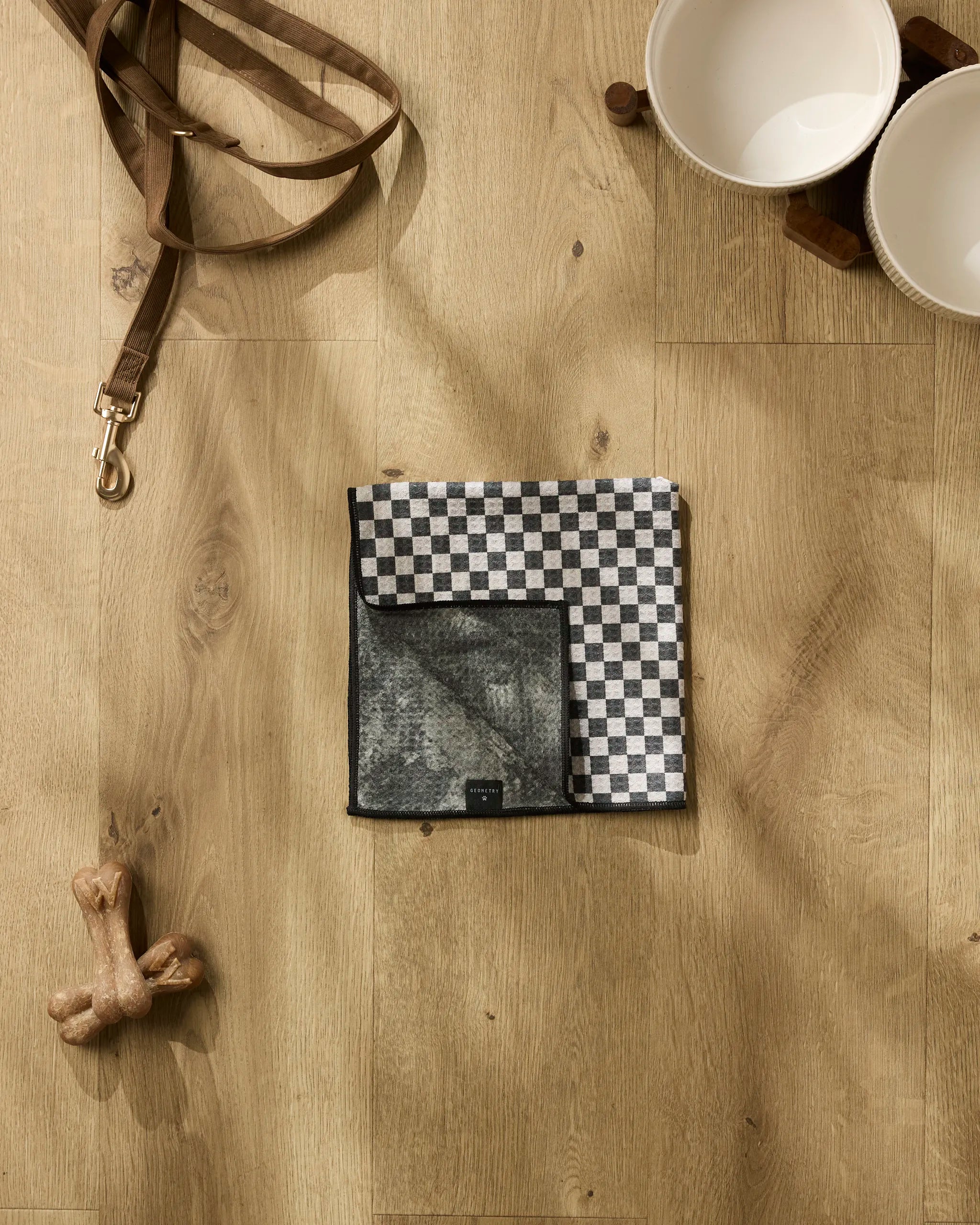 A flat lay photo of Cool Pup dog accessories by Geometry on a wooden floor, featuring a brown leash, two white bowls on a wooden stand, a black-and-white checked bandana, dog bones, and an ultra-absorbent dog towel.