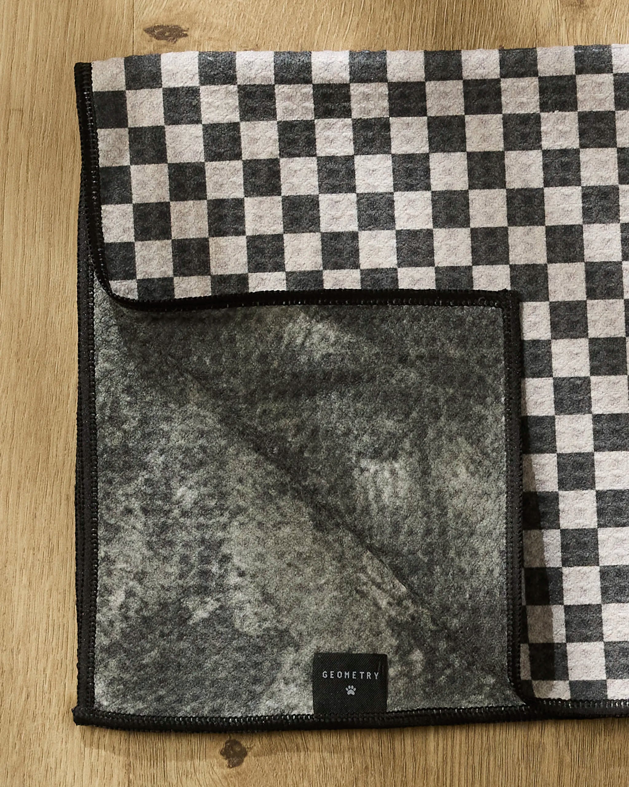 A black and white checkered Cool Pup towel by Geometry rests on a wooden surface with a corner folded back, showing its textured gray underside and a black Geometry label.