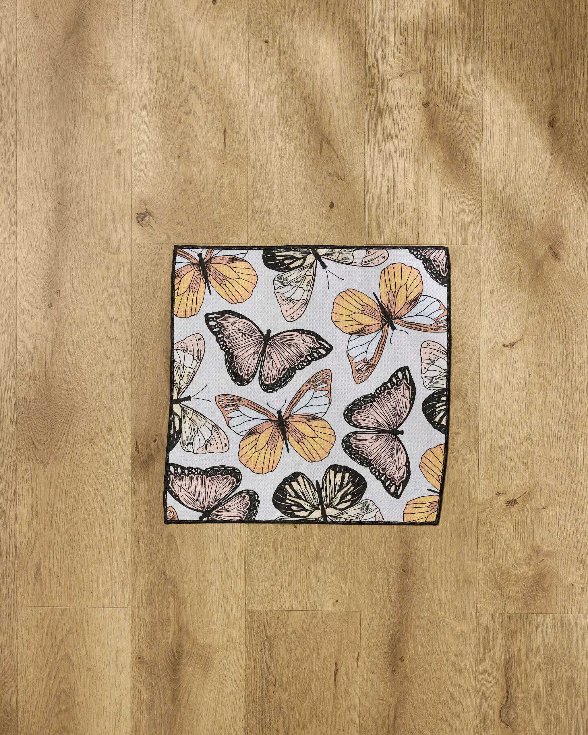The Geometry "Cute Butterflies" square mat, made from recycled pet towel material and featuring orange, yellow, black, and white butterflies, is displayed on a light wood floor with visible grain.