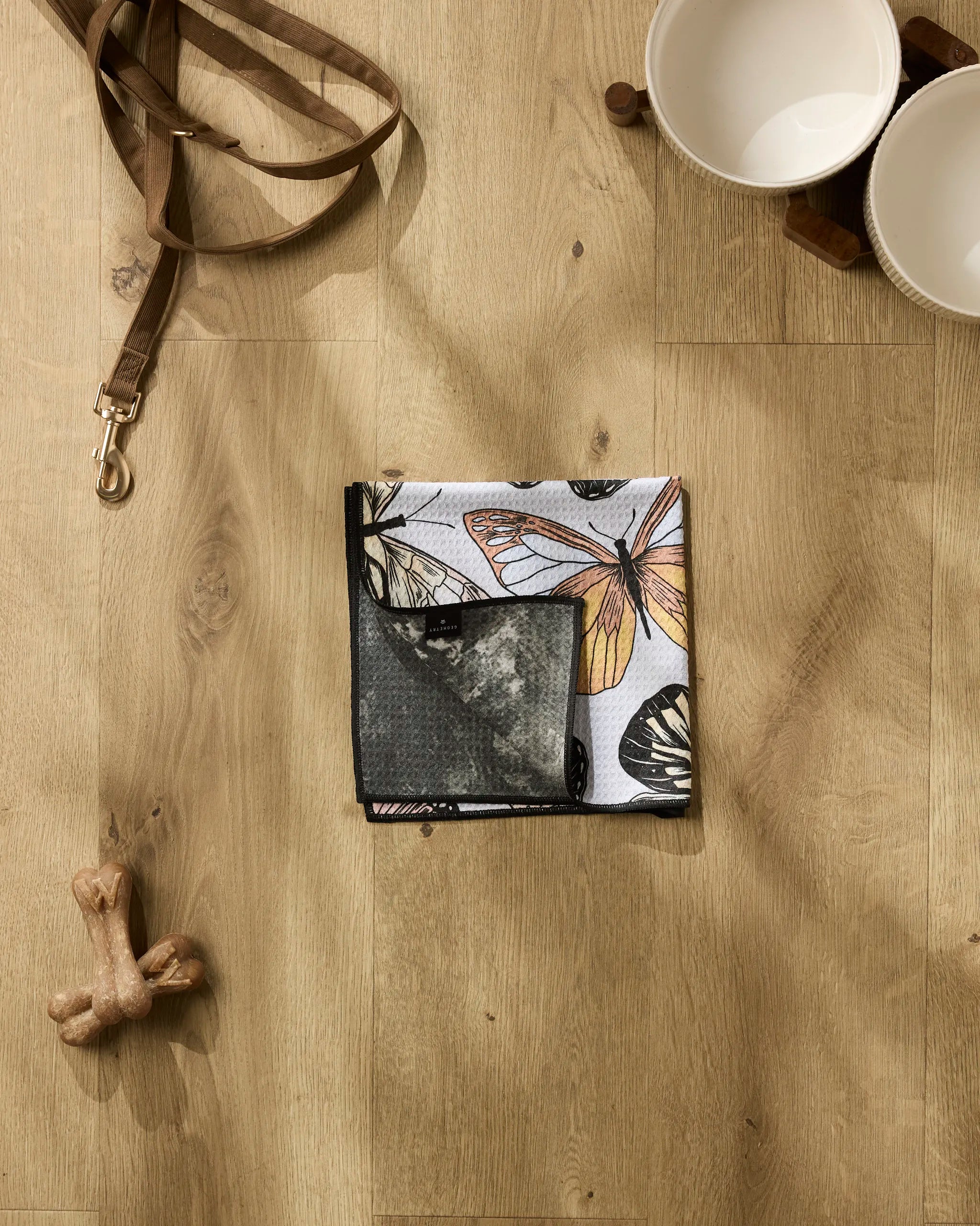 A folded Geometry "Cute Butterflies" bandana sits on a wooden floor beside a brown leash, ceramic dog bowls, bone treats, and an ultra-absorbent dog towel.