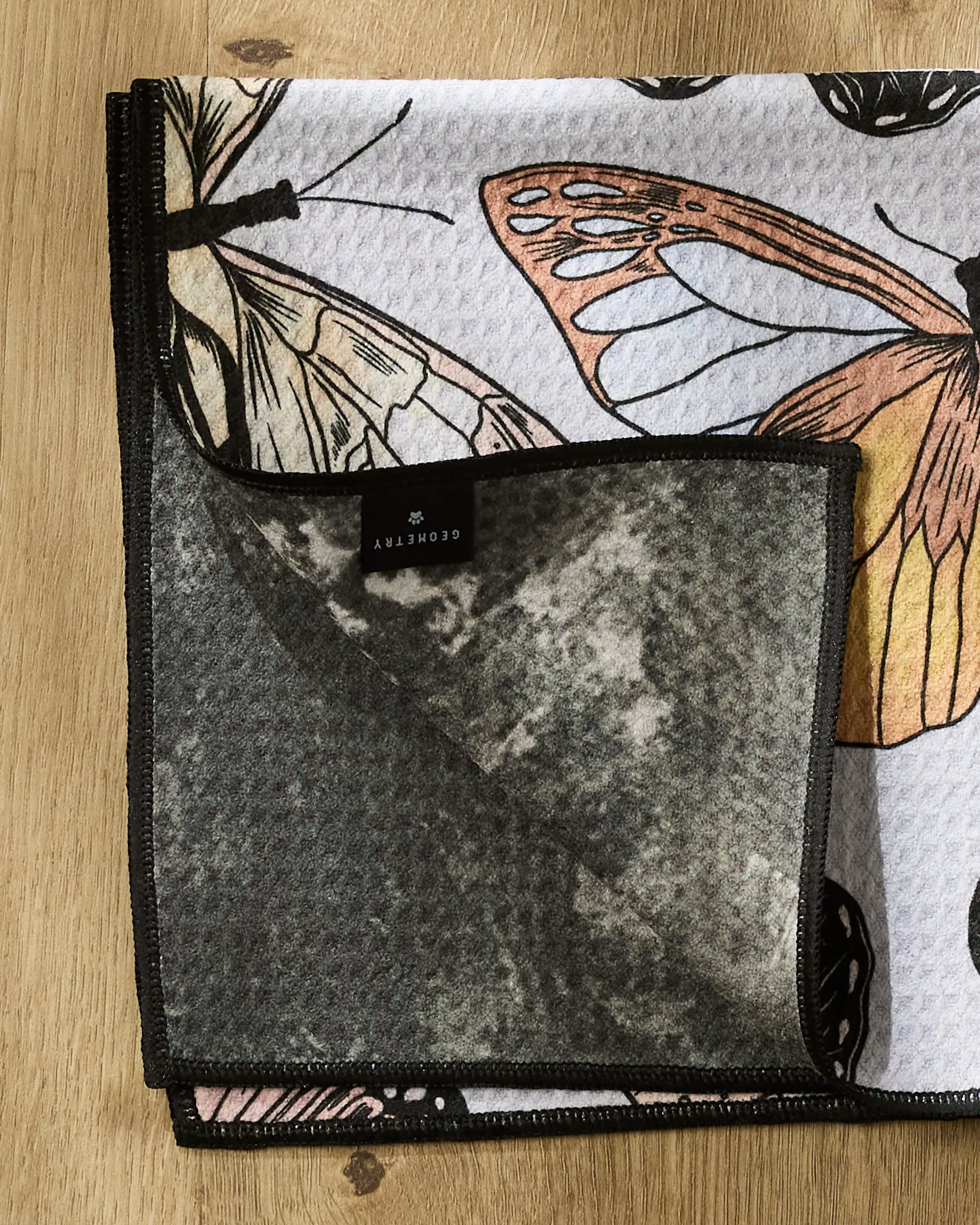 A soft "Cute Butterflies" Paw towel by Geometry rests on a wooden surface, partially folded to reveal its cozy gray fleece underside and the visible brand label.