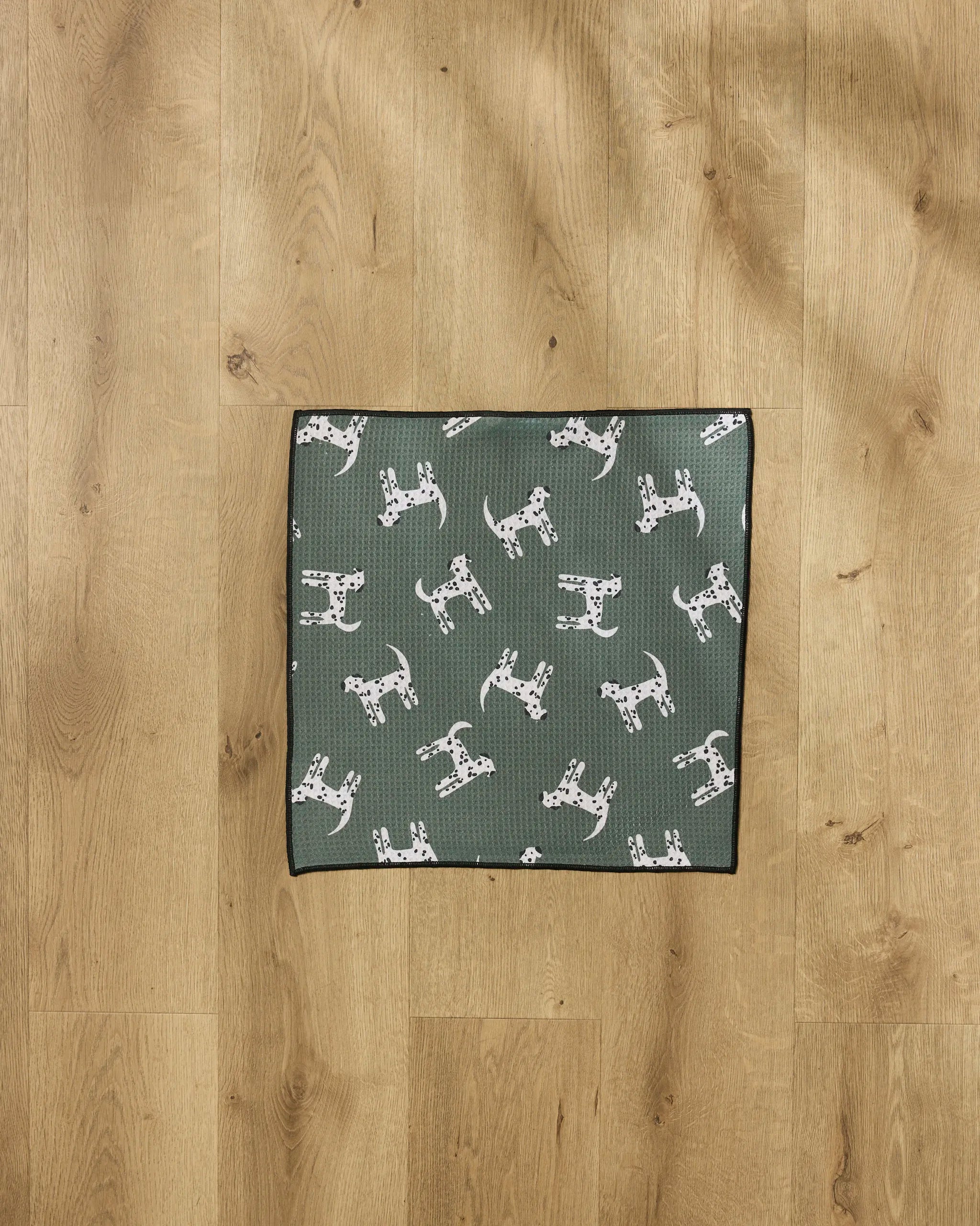 The Coit Creative "Dalmation Dogs" recycled PET towel, featuring a white and black spotted dog pattern, is laid flat on a light wood floor.
