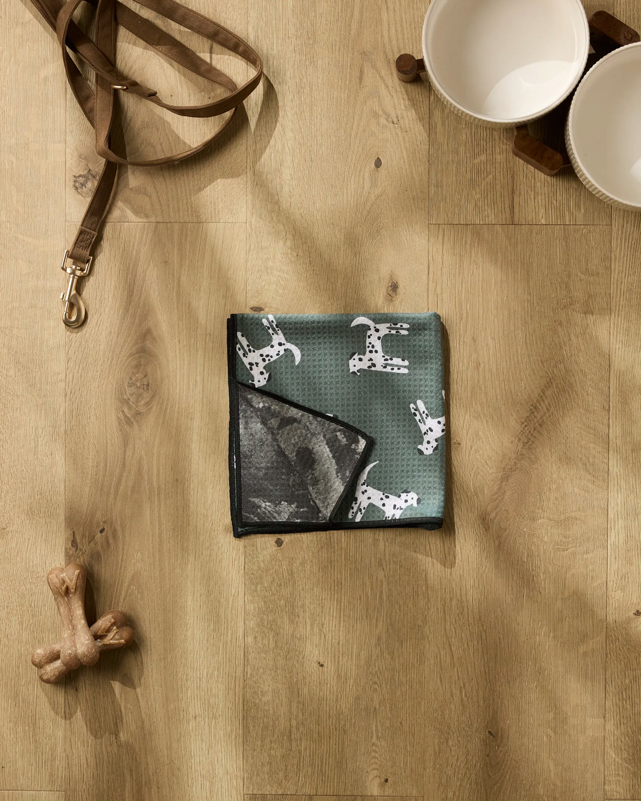 A flat lay of Coit Creative’s Dalmation Dogs accessories on a wooden floor, including a brown leash, white bowls on a stand, Dalmatian print bandana, bone-shaped chew toy, and an ultra-absorbent dog towel for post-walk drying.