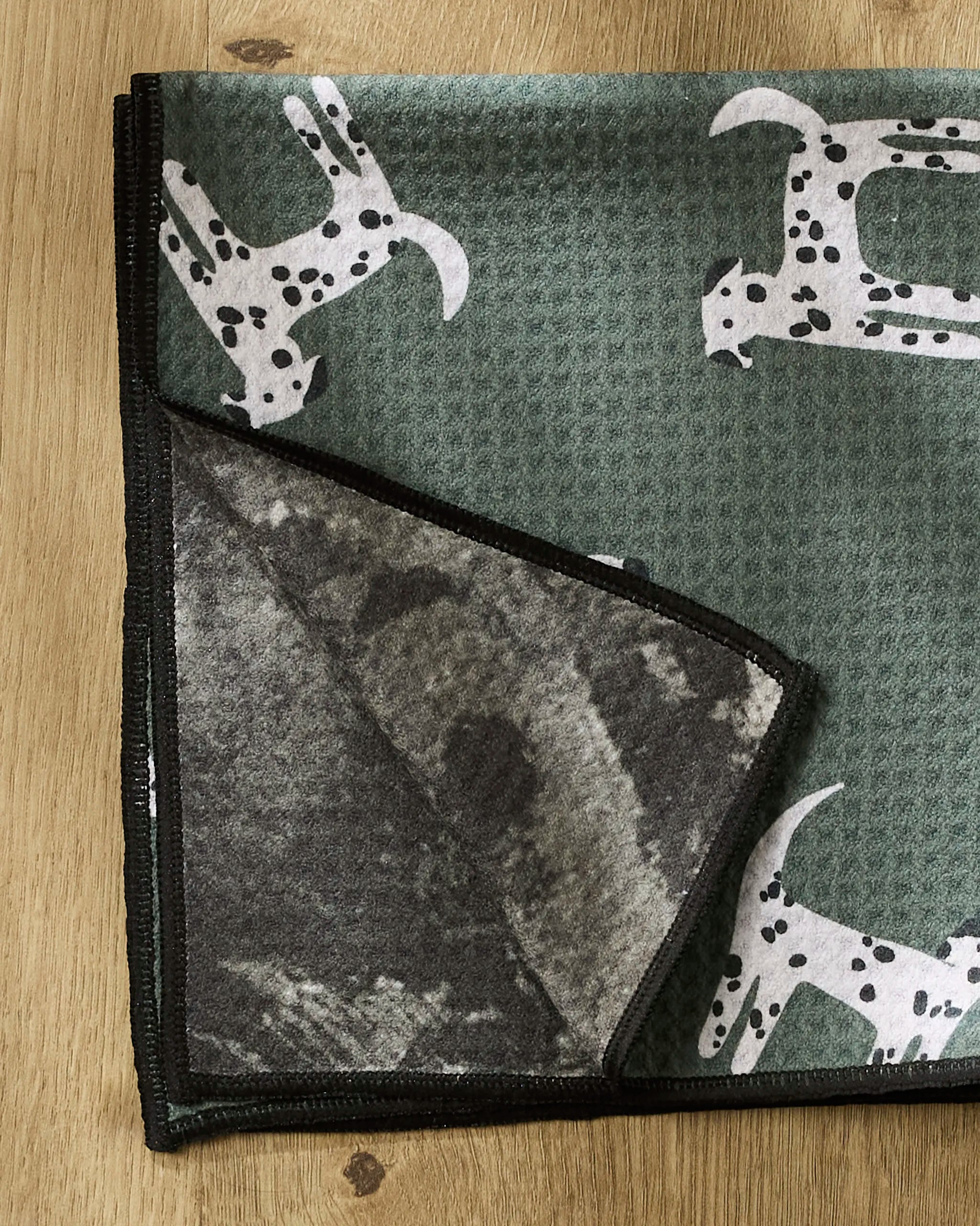 The Coit Creative “Dalmatian Dogs” towel is a green, ultra-absorbent recycled pet towel with white cartoon dalmatians, a black border, and dark gray underside, shown flipped at one corner on a wooden surface.
