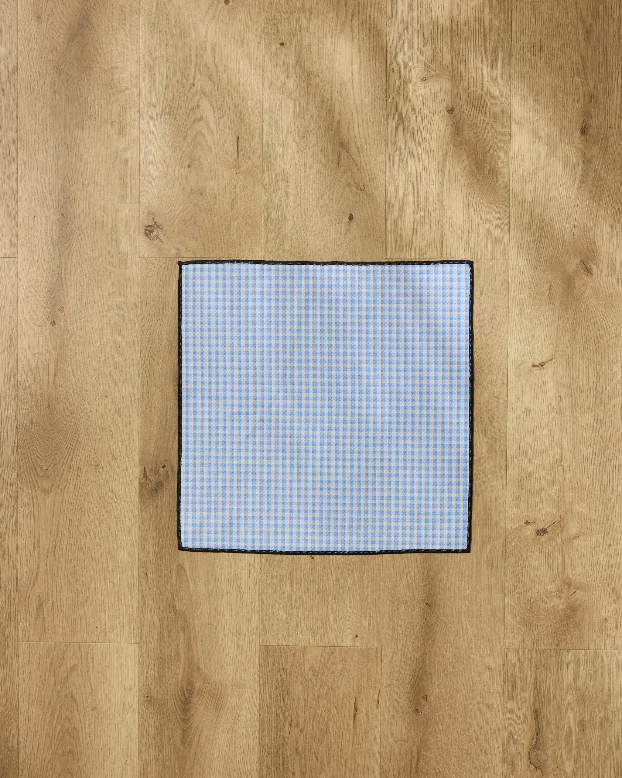 The Geometry Dog Delight towel, featuring blue and white checks, a black border, and ultra-absorbent material, is laid flat on a light wood floor.