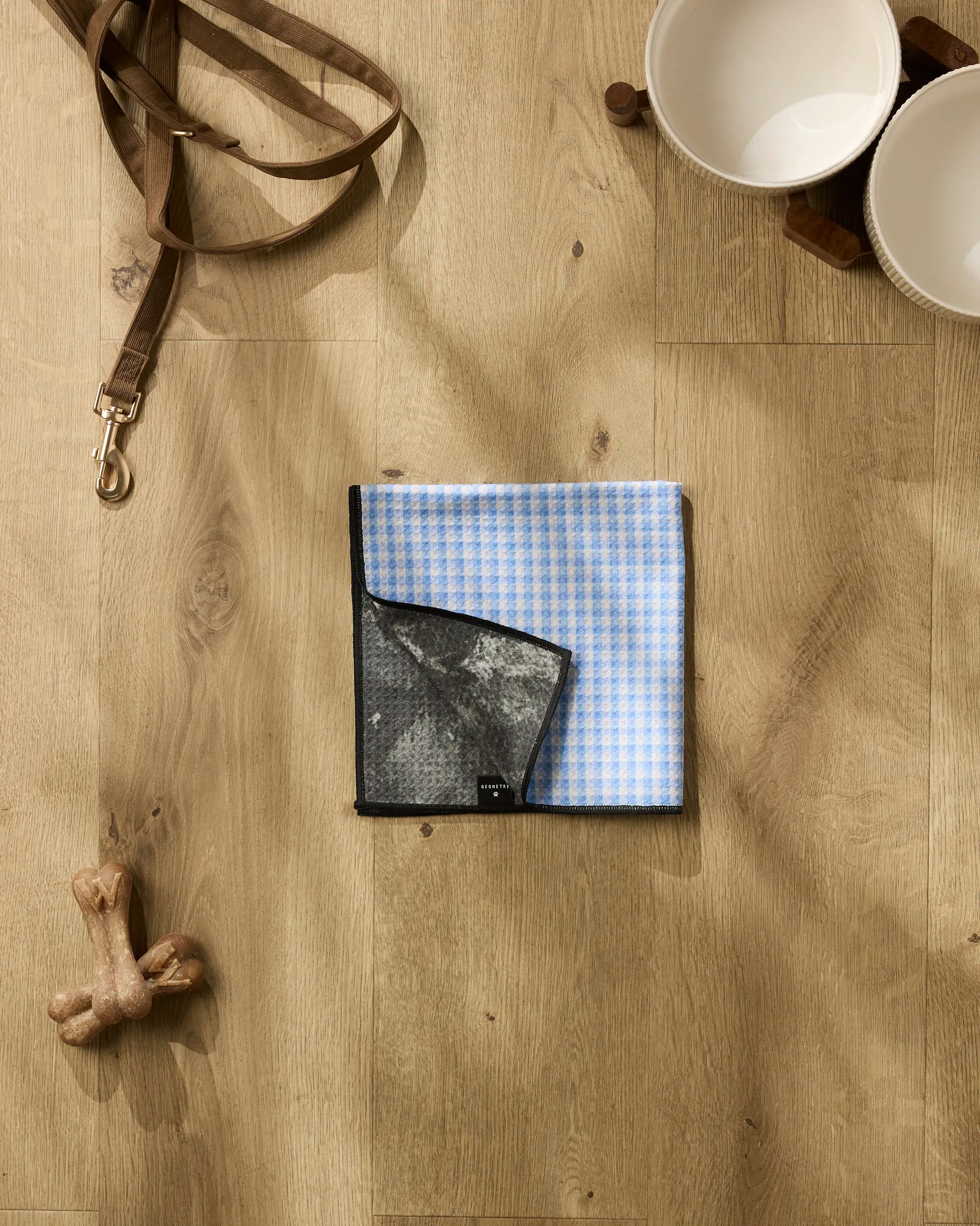 The Geometry Dog Delight features a blue and white checkered bandana, a leash, two white bowls on a stand, a bone-shaped chew toy, and an ultra-absorbent dog towel—perfect for quick cleanups after playtime.