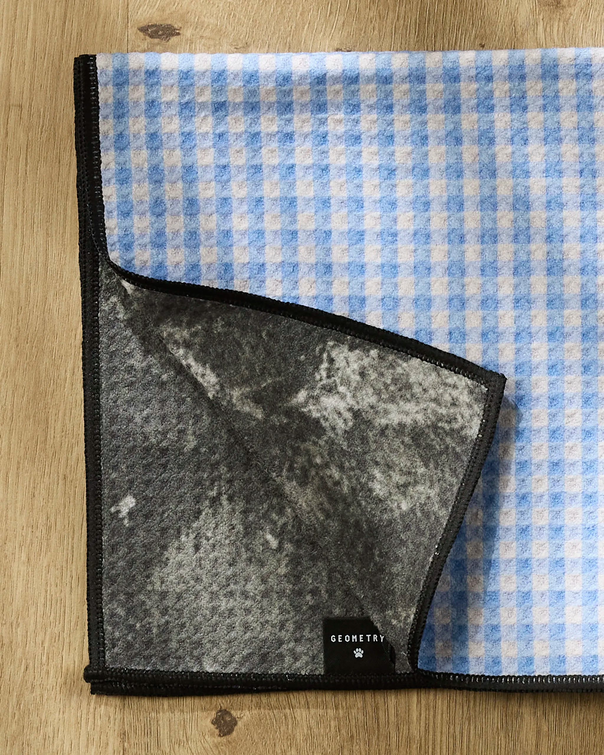 A blue and white checkered Dog Delight towel by Geometry rests on a wooden surface, one corner flipped to reveal a gray and black underside and a small "GEOMETRY" tag. This ultra-absorbent towel is ideal for drying your pet after messy adventures.