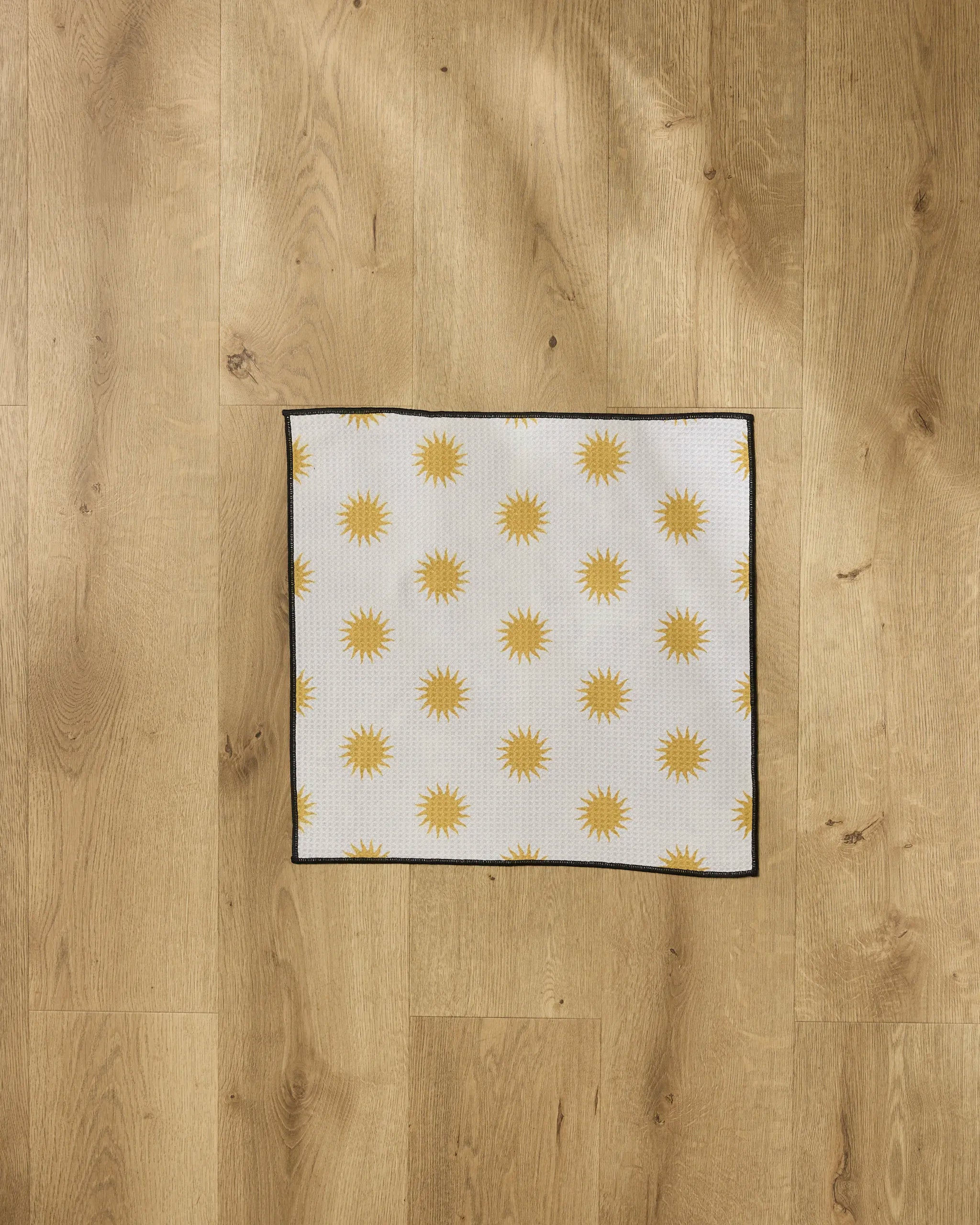 The Geometry Feeling Sunny paw towel, featuring a square shape with a repeating yellow sun pattern, black border, and white background, is laid flat on a light-colored wooden floor.
