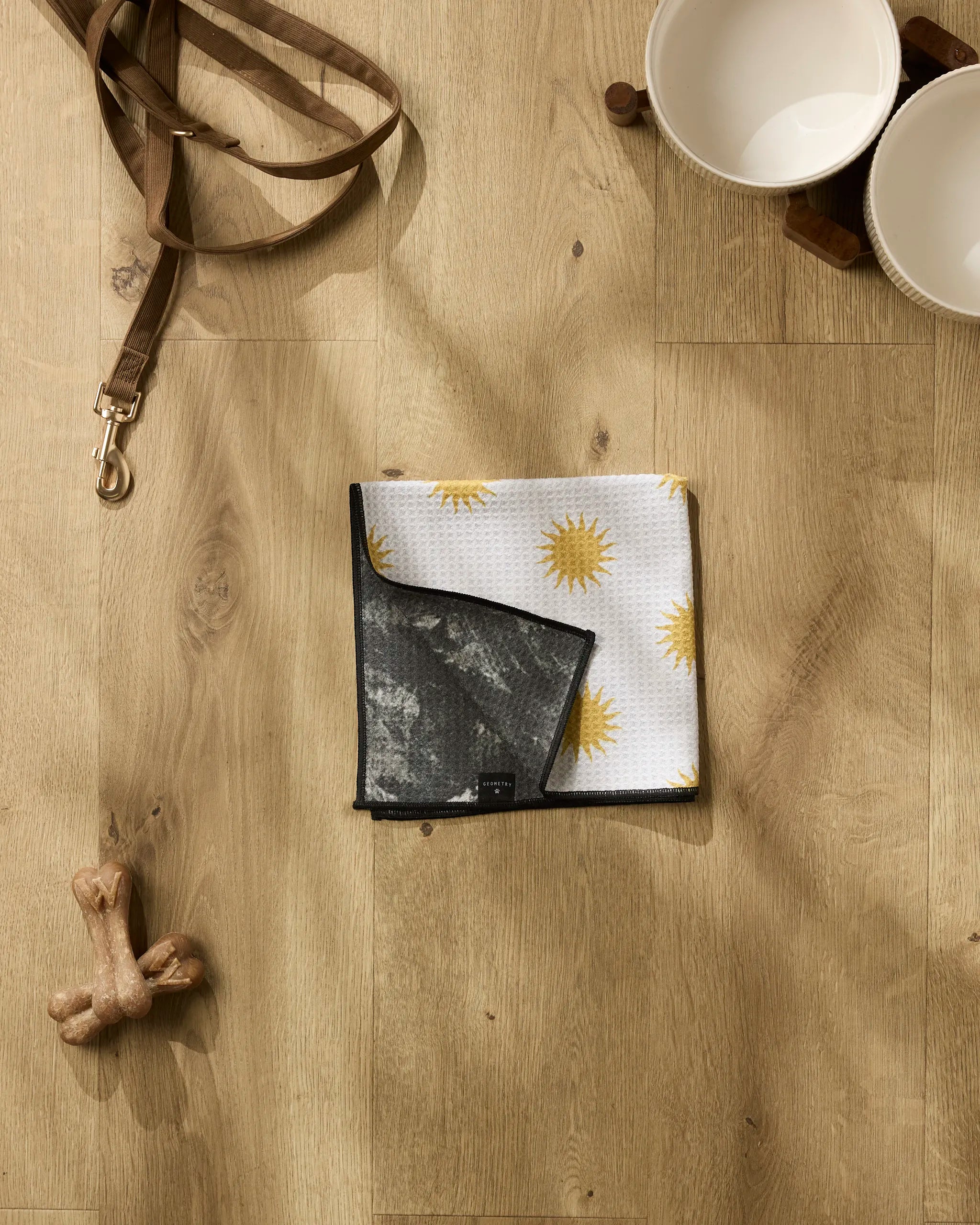 Geometry’s Feeling Sunny set—featuring a tan dog leash, two white pet bowls on a wood stand, a recycled towel with yellow sun patterns, and a dog bone toy—is arranged on a light wood floor.